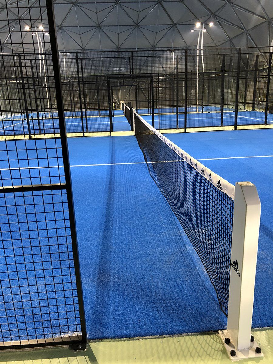 Campo da padel con superficie e rete blu, all'interno di una grande struttura coperta.