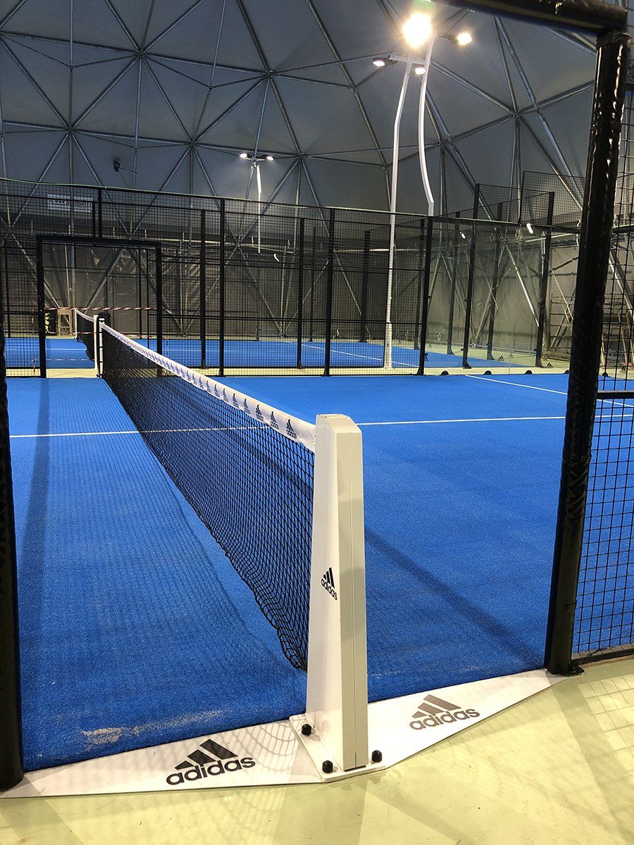 Campo da padel blu con rete, pali bianchi, recinzione nera e loghi Adidas; ambiente al coperto.
