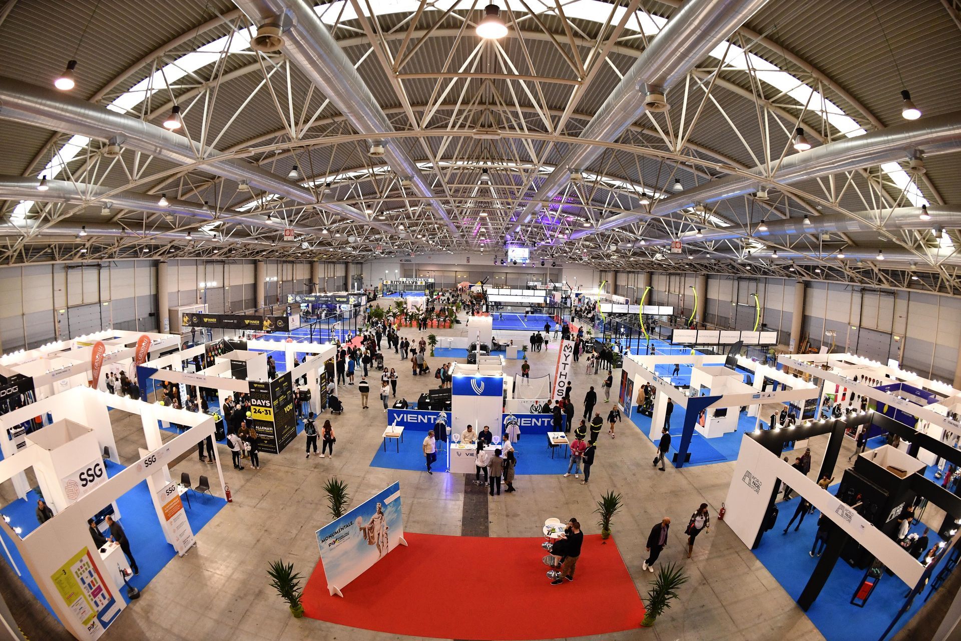 Ampia sala congressi piena di stand di venditori e partecipanti; espositori blu e bianchi, area con tappeto rosso.
