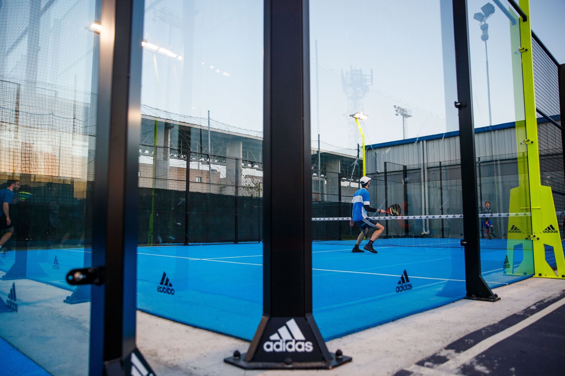 Campo da padel con superficie e giocatori blu, visibile attraverso pannelli di vetro. Pali e rete neri e cielo azzurro.