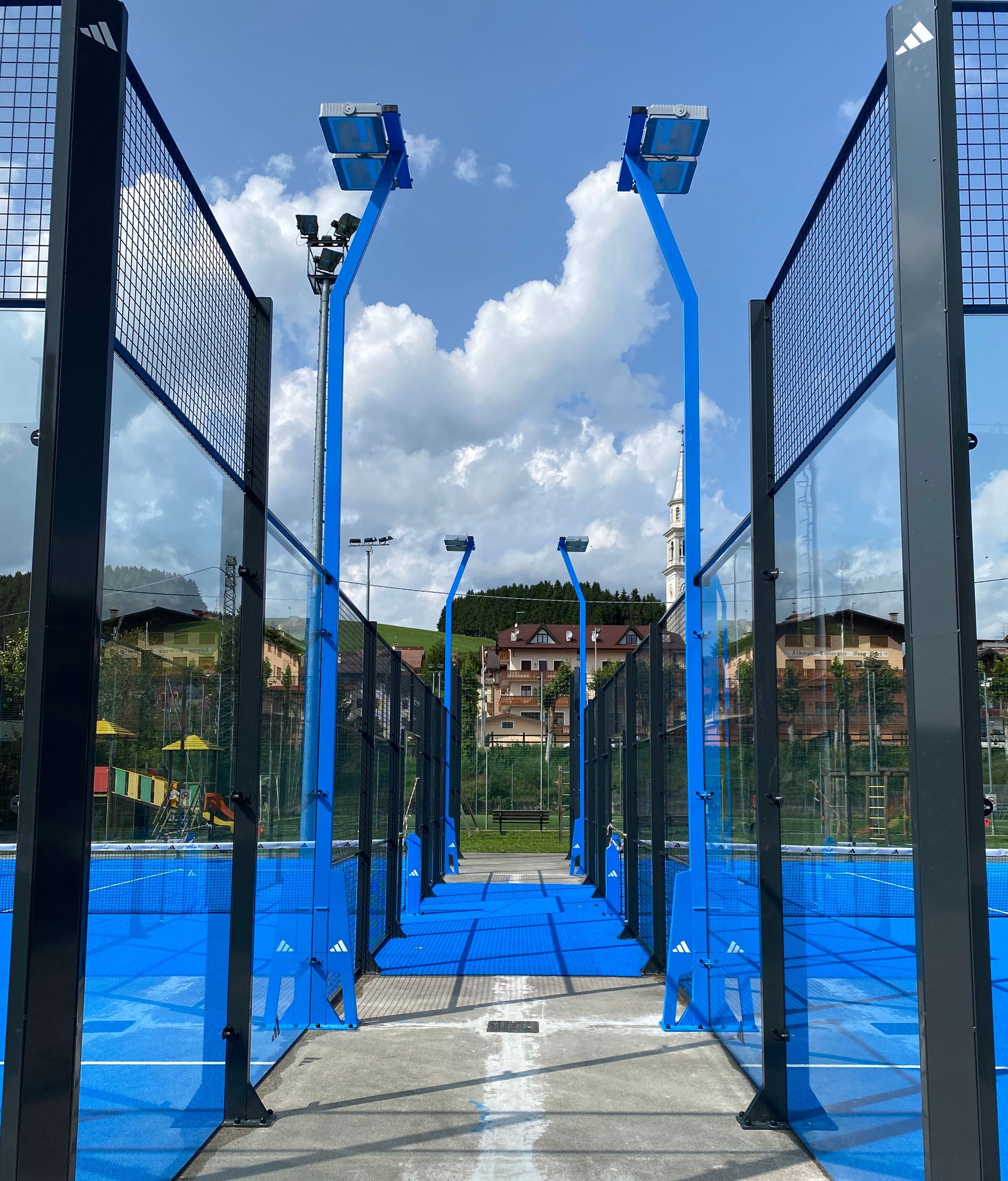 Campo da padel blu con pareti in vetro, pavimento blu e illuminazione, sullo sfondo di un cielo soleggiato 