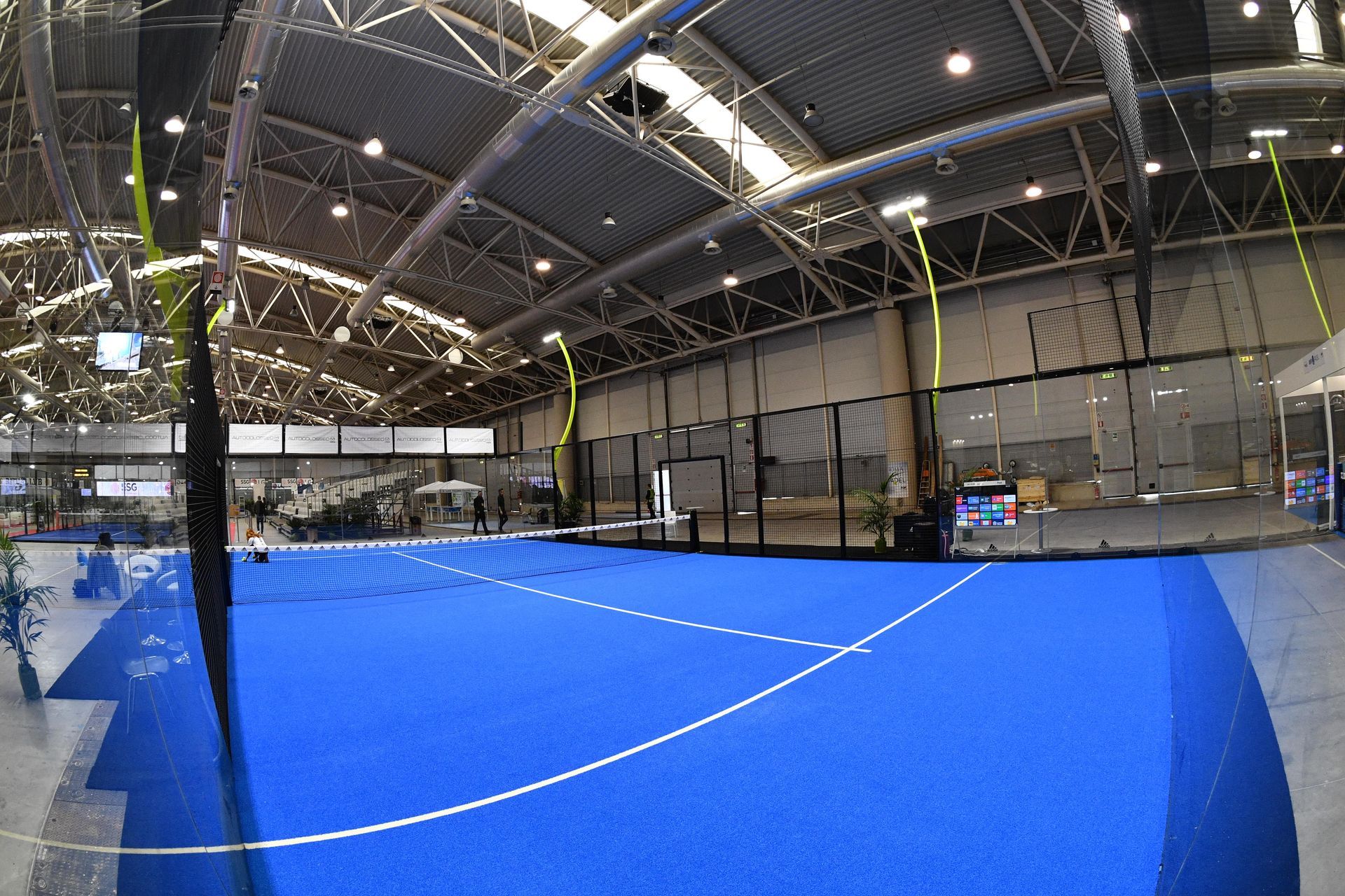 Campo da padel indoor con superficie blu e linee bianche, reti e illuminazione dall'alto.