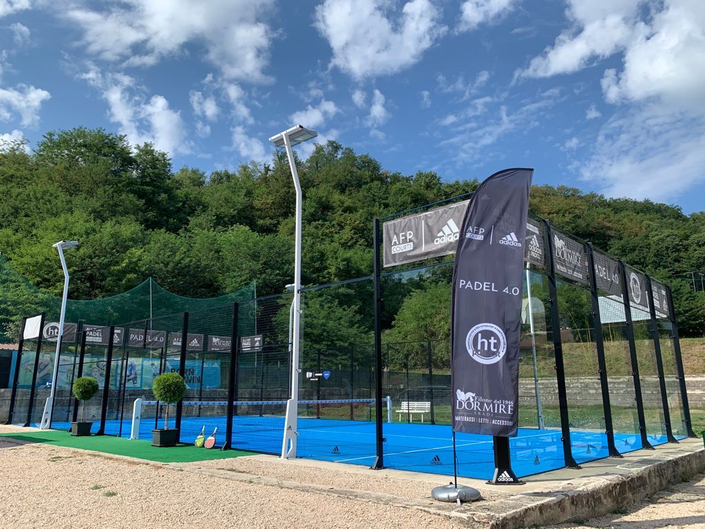 Campo da padel blu all'aperto con rete, recinzioni perimetrali e bandiere degli sponsor sotto un cielo azzurro.