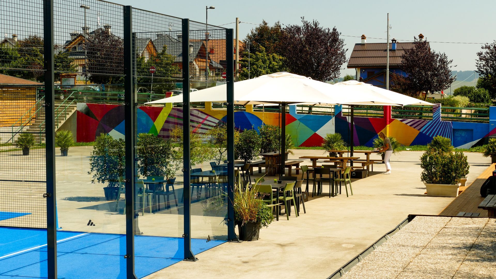 Bar all'aperto accanto a un campo sportivo blu. Tavoli e ombrelloni su un patio in cemento. Muro colorato sullo sfondo.