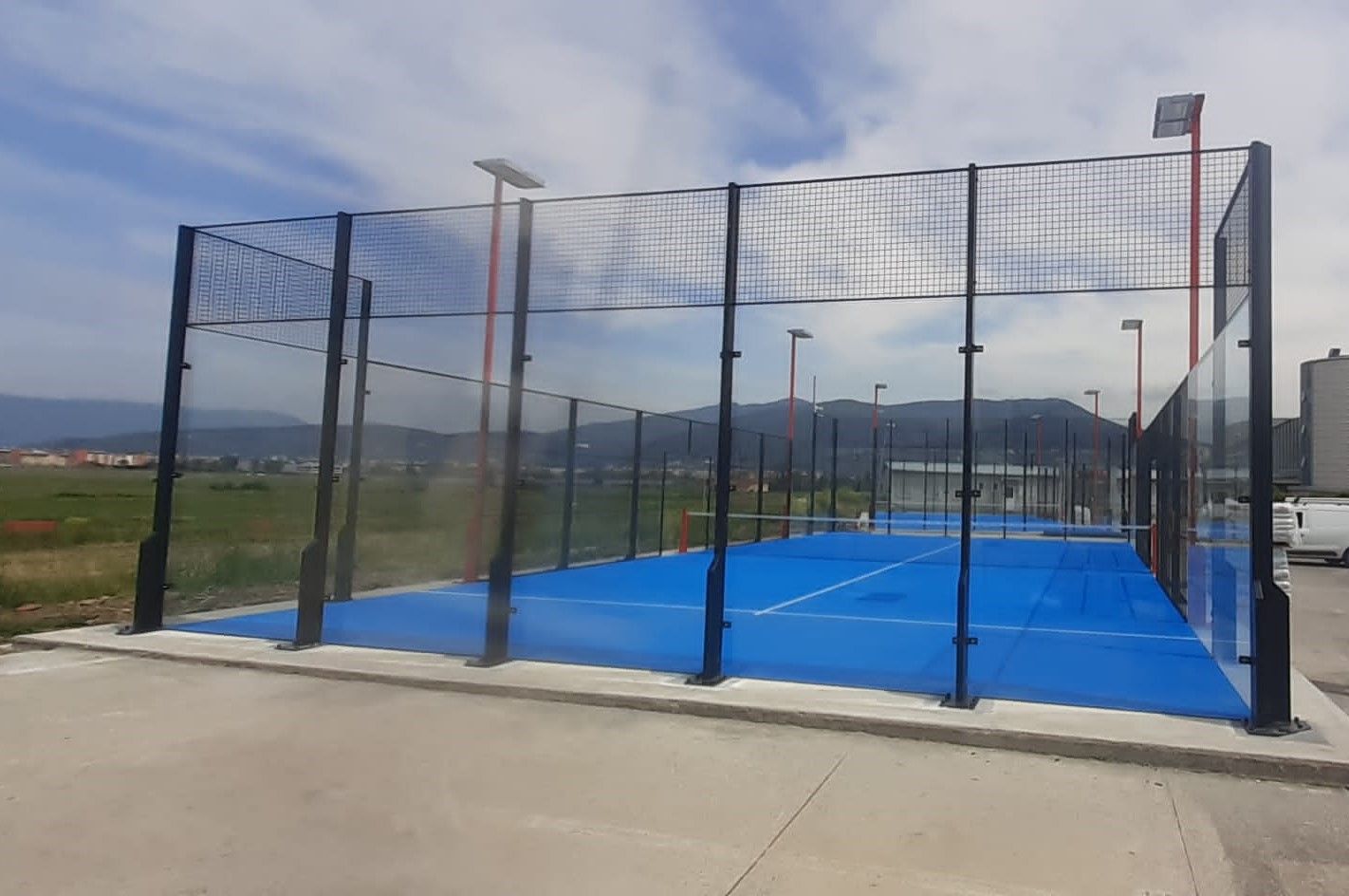 Campo da padel blu recintato da una recinzione metallica, sotto un cielo parzialmente nuvoloso.