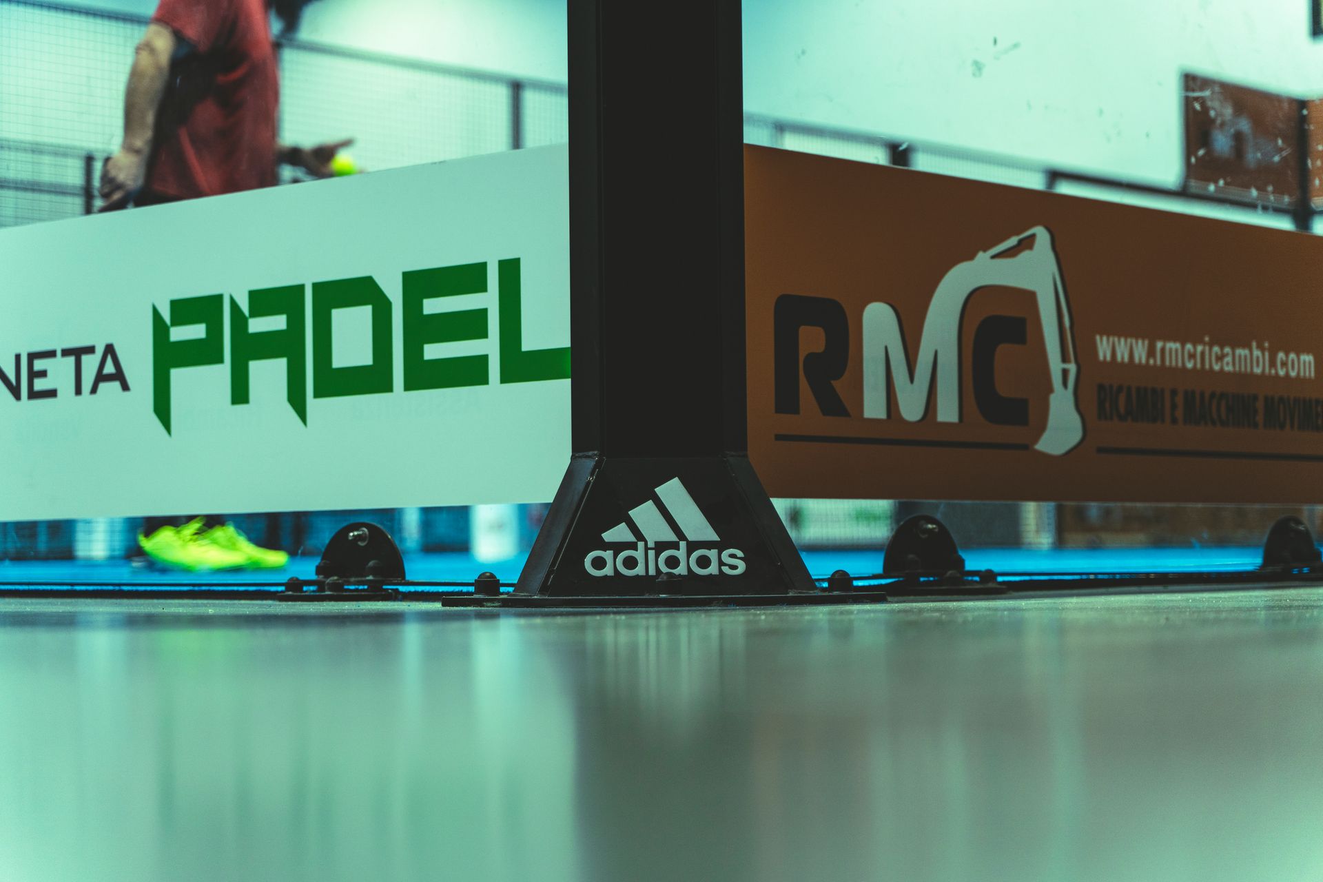 Rete da padel con marchio Adidas sul campo blu, cartelli degli sponsor verdi e arancioni sullo sfondo.