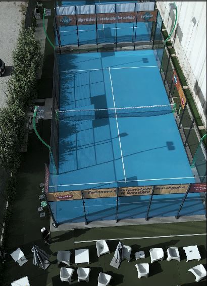 Vista dall'alto di un campo da padel blu con rete, circondato da recinzione, verde e sedie bianche.