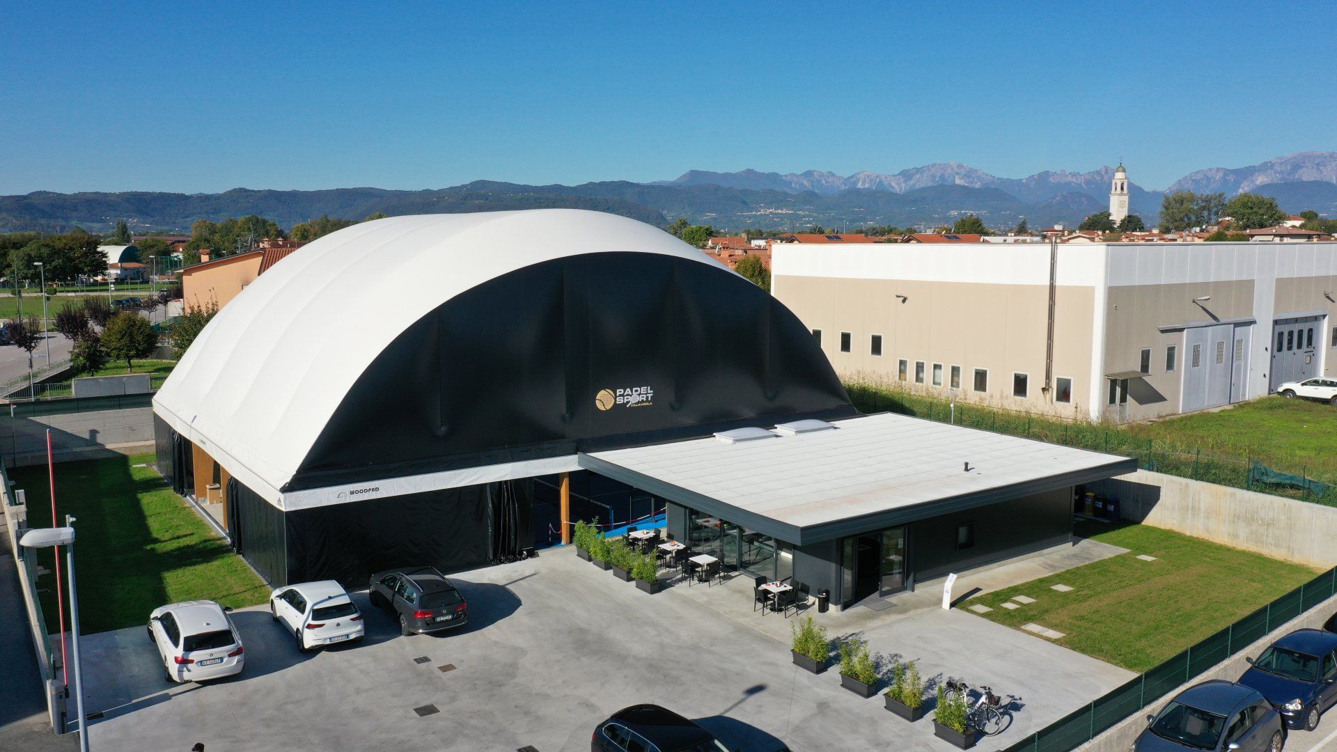 Vista esterna di un grande edificio con tetto curvo e facciata nera, a cui si aggiunge un moderno edificio annesso. 