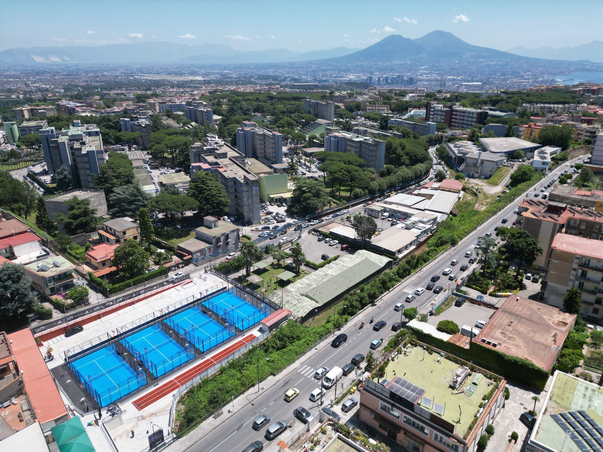 Vista aerea di una città con campi da paddle tennis blu in primo piano, una strada con auto e una montagna sullo sfondo.