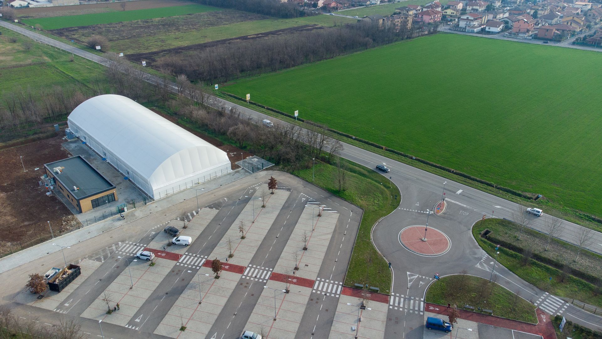 Vista aerea di un grande edificio bianco con tende, parcheggio, strada e campi verdi.