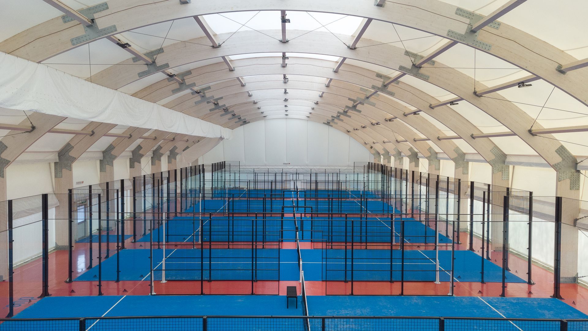 Campo da padel tennis al coperto con campi blu, pareti in vetro e soffitto a volta.