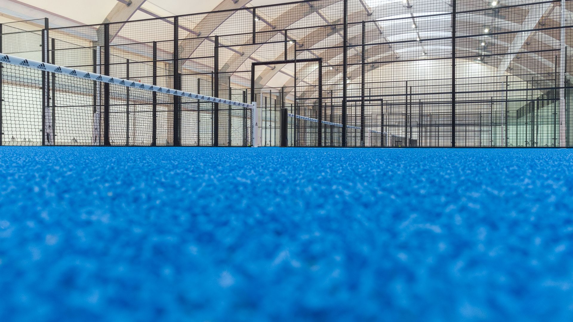 Campo da padel blu con rete, circondato da pareti in vetro e soffitto con struttura metallica.