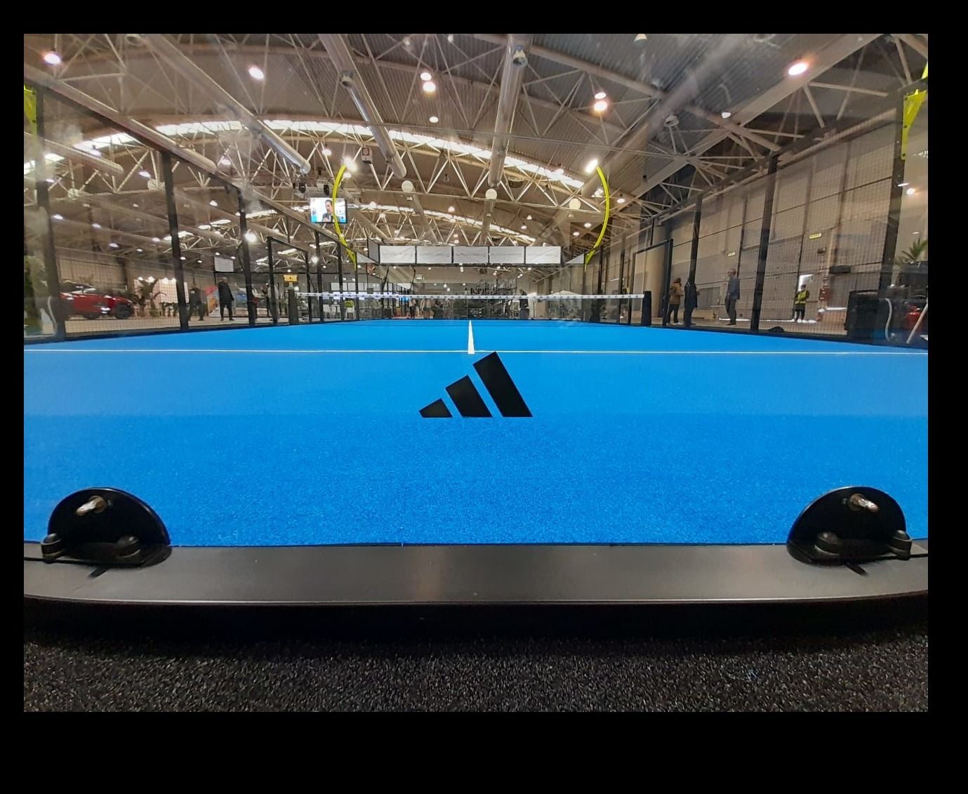 Campo da padel indoor blu con logo Adidas; reti nere, giocatori visibili.