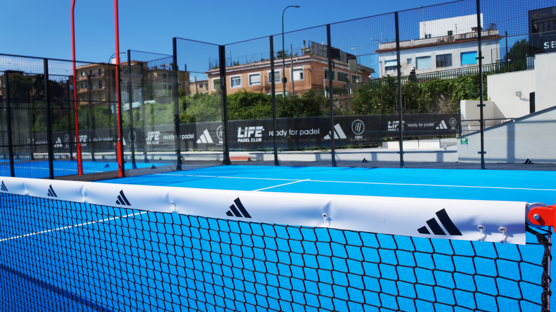 Campo da padel blu con rete e marchio Adidas. Edifici sullo sfondo.