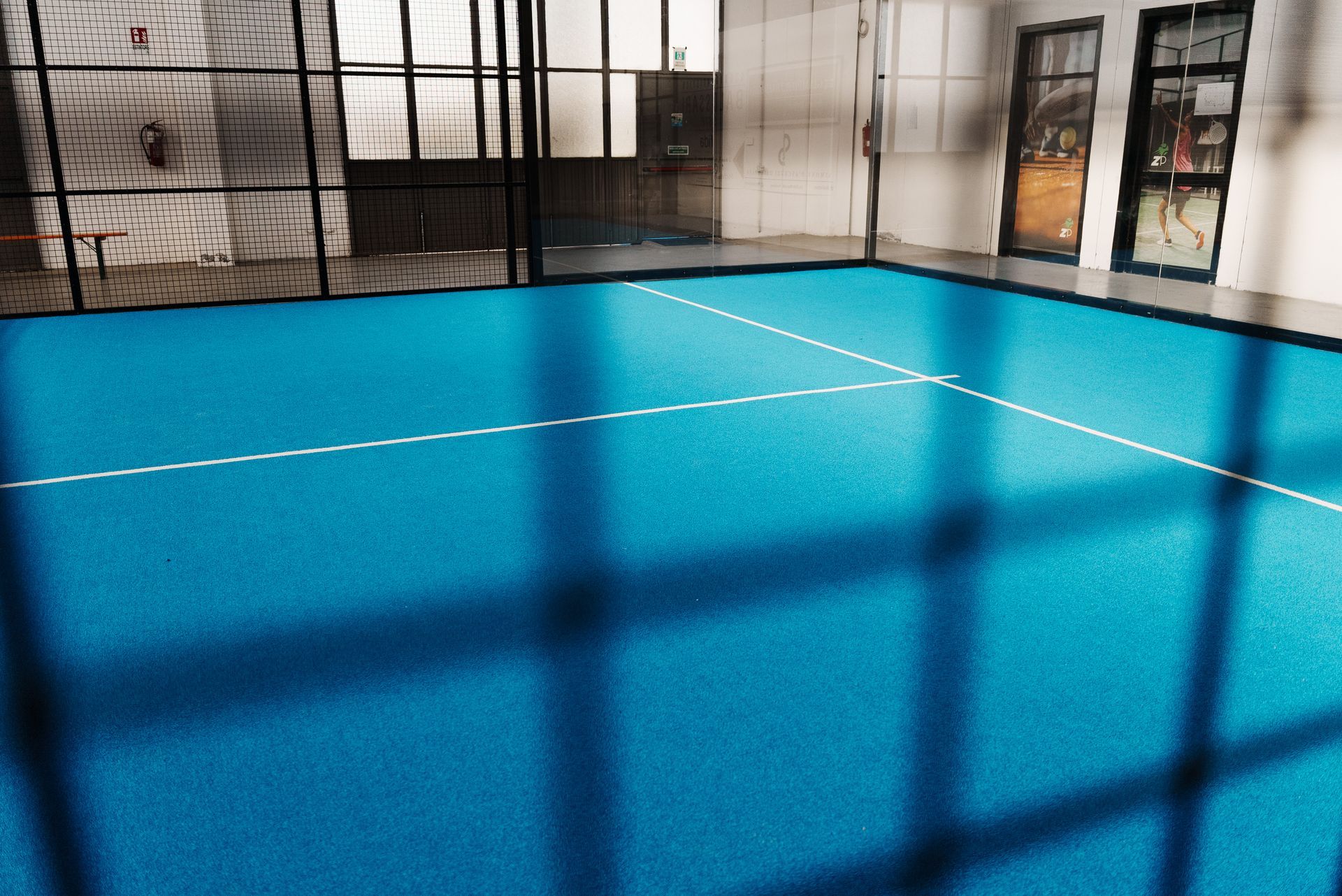 Campo da padel coperto blu con linee bianche, pareti in vetro e rete nera.