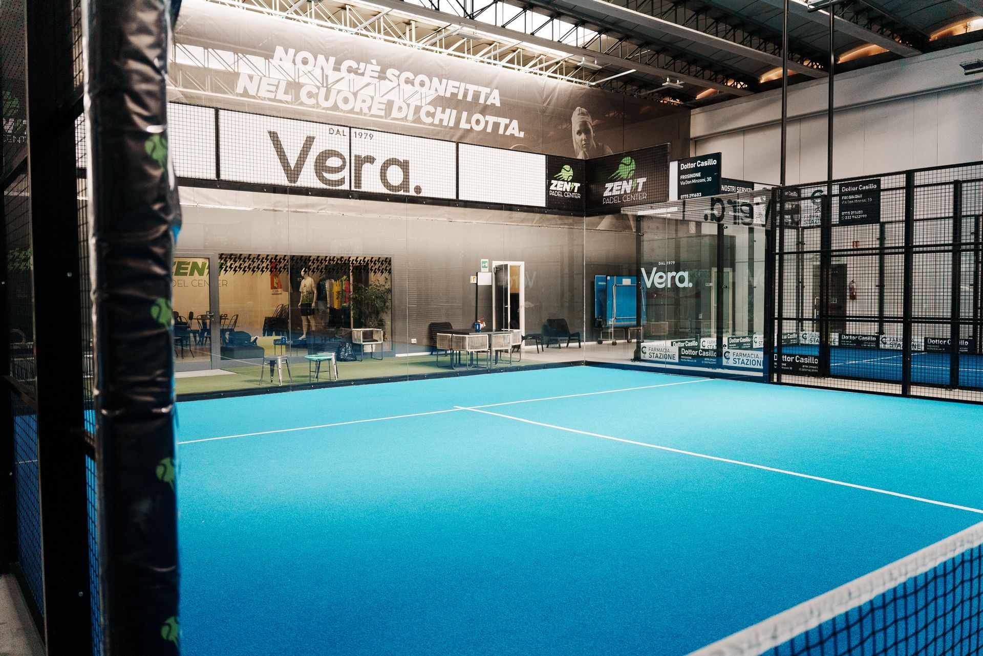 Campo da padel al coperto, superficie blu, recinzione nera, insegna Vera, illuminazione dall'alto.