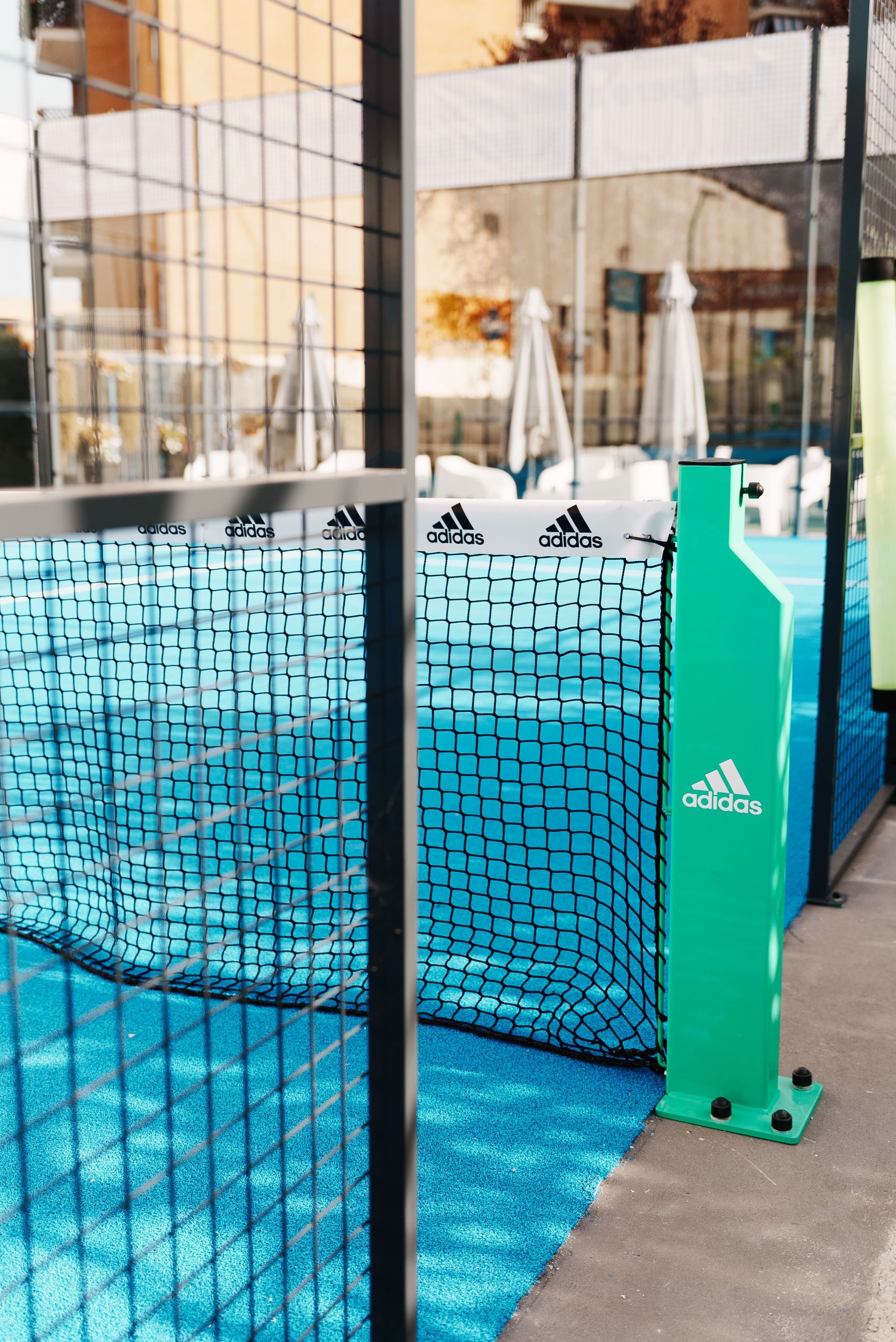 Campo da padel Adidas verde e blu con rete, logo e ombrelloni sullo sfondo.