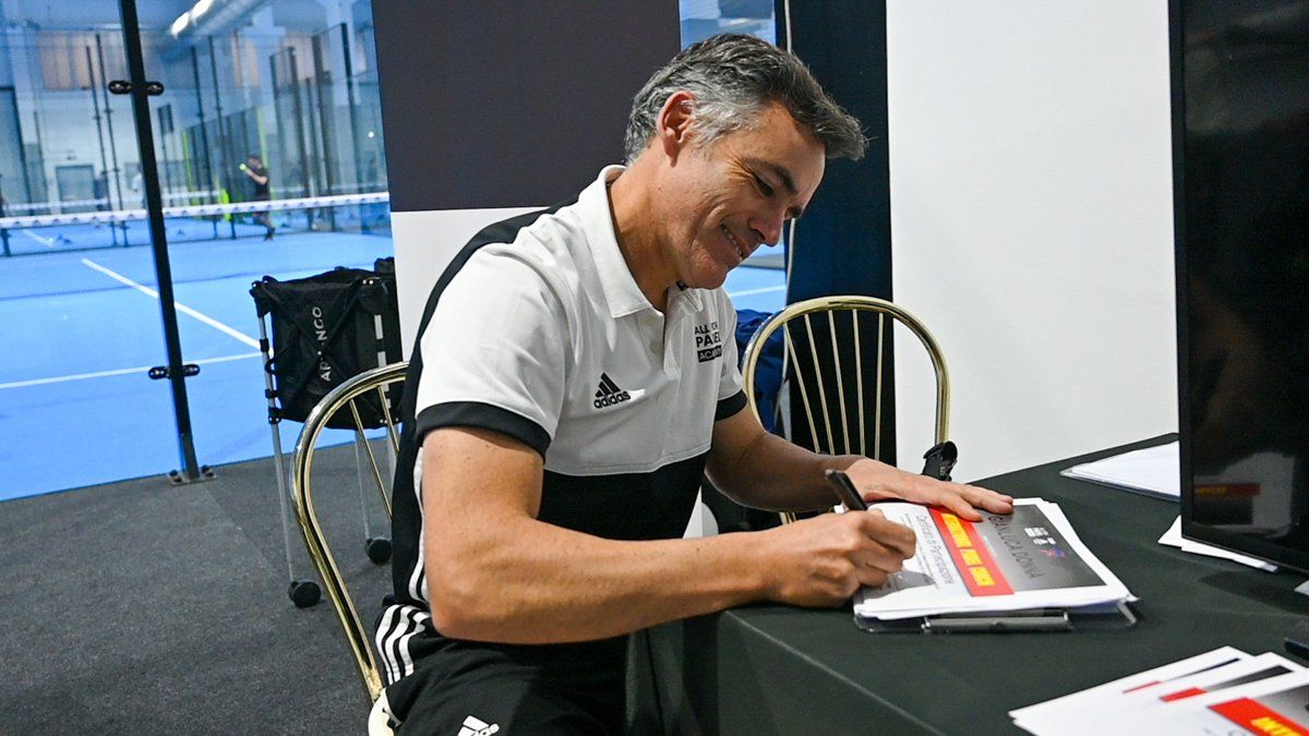 Uomo in camicia bianca che firma documenti alla scrivania, ambiente interno, campo da paddle tennis sullo sfondo.