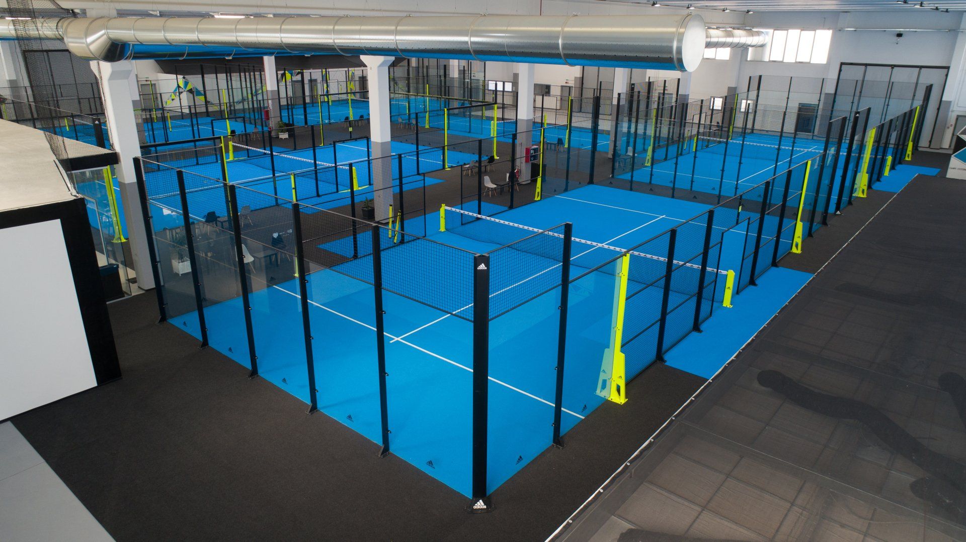 Campo da padel indoor con più campi blu, bordi neri e travi di sostegno.