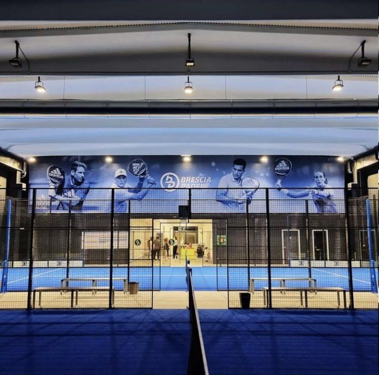 Campo da padel al coperto, superficie blu, sagome dei giocatori sul muro, scuro.