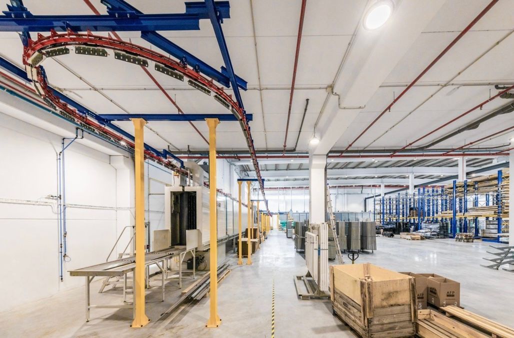 Interno di una fabbrica con trasportatore aereo e scatole a terra. Colori blu, giallo e rosso.