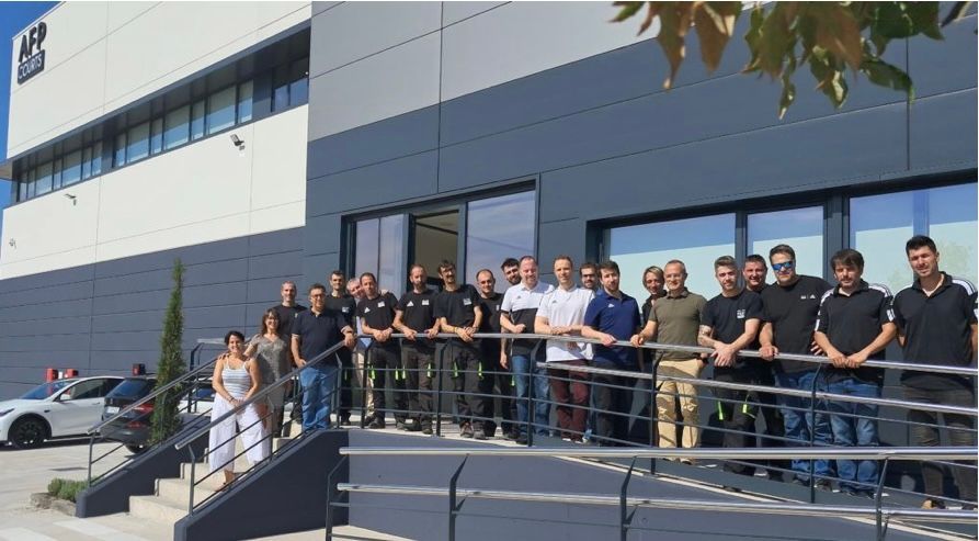 Gruppo di persone in piedi fuori da un edificio grigio con un cartello; giornata di sole.