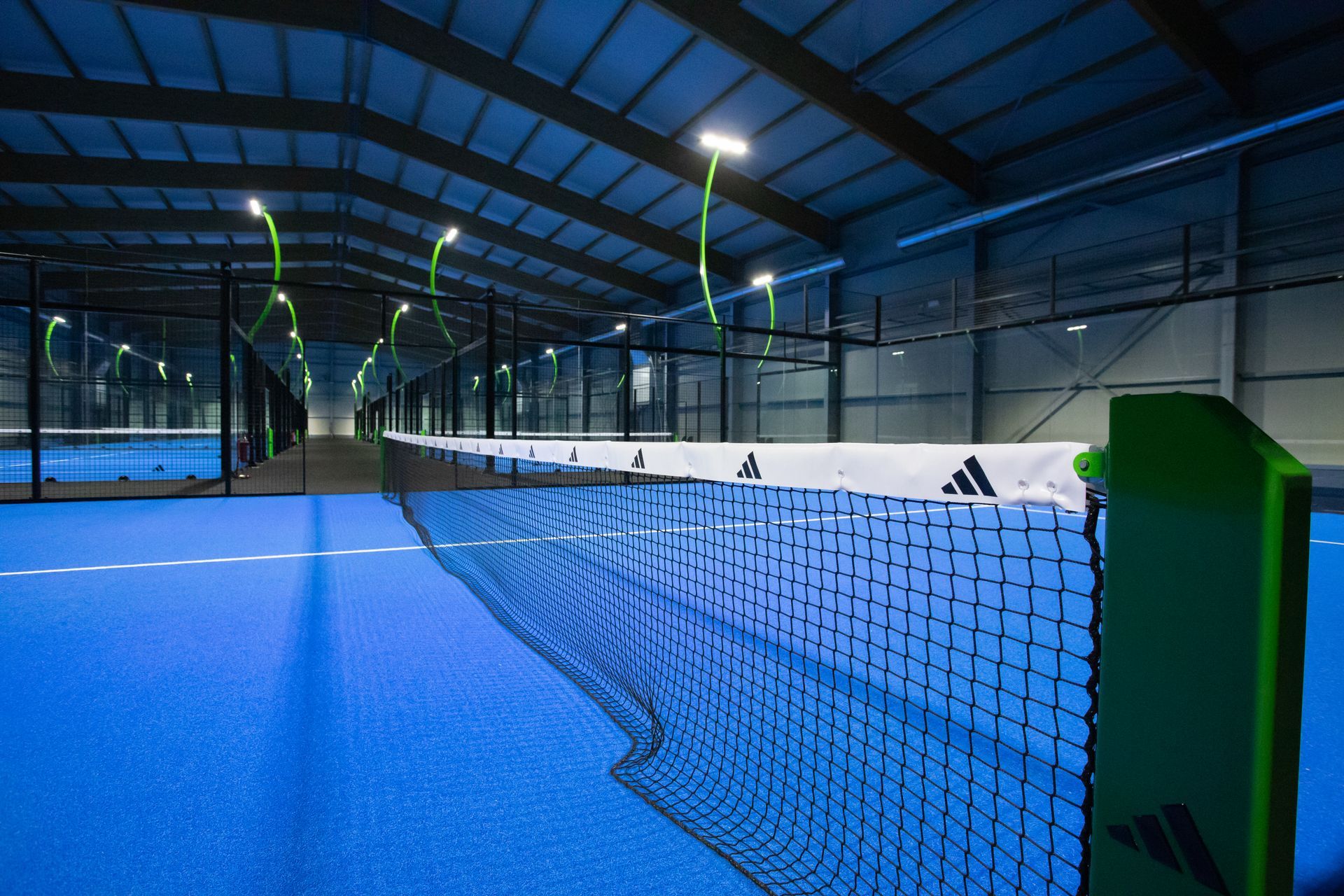 Campo da padel indoor, superficie blu, rete nera, marchio Adidas, dettagli verdi, illuminazione dall'alto.