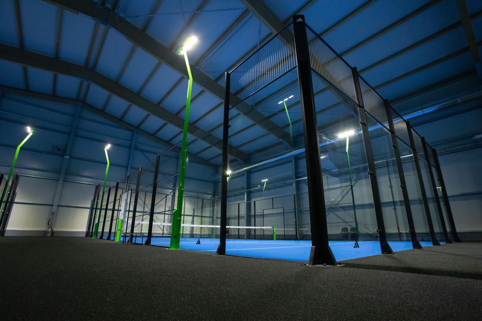 Campo da padel indoor, pavimento blu, rete e supporti neri, illuminazione verde, soffitto alto.