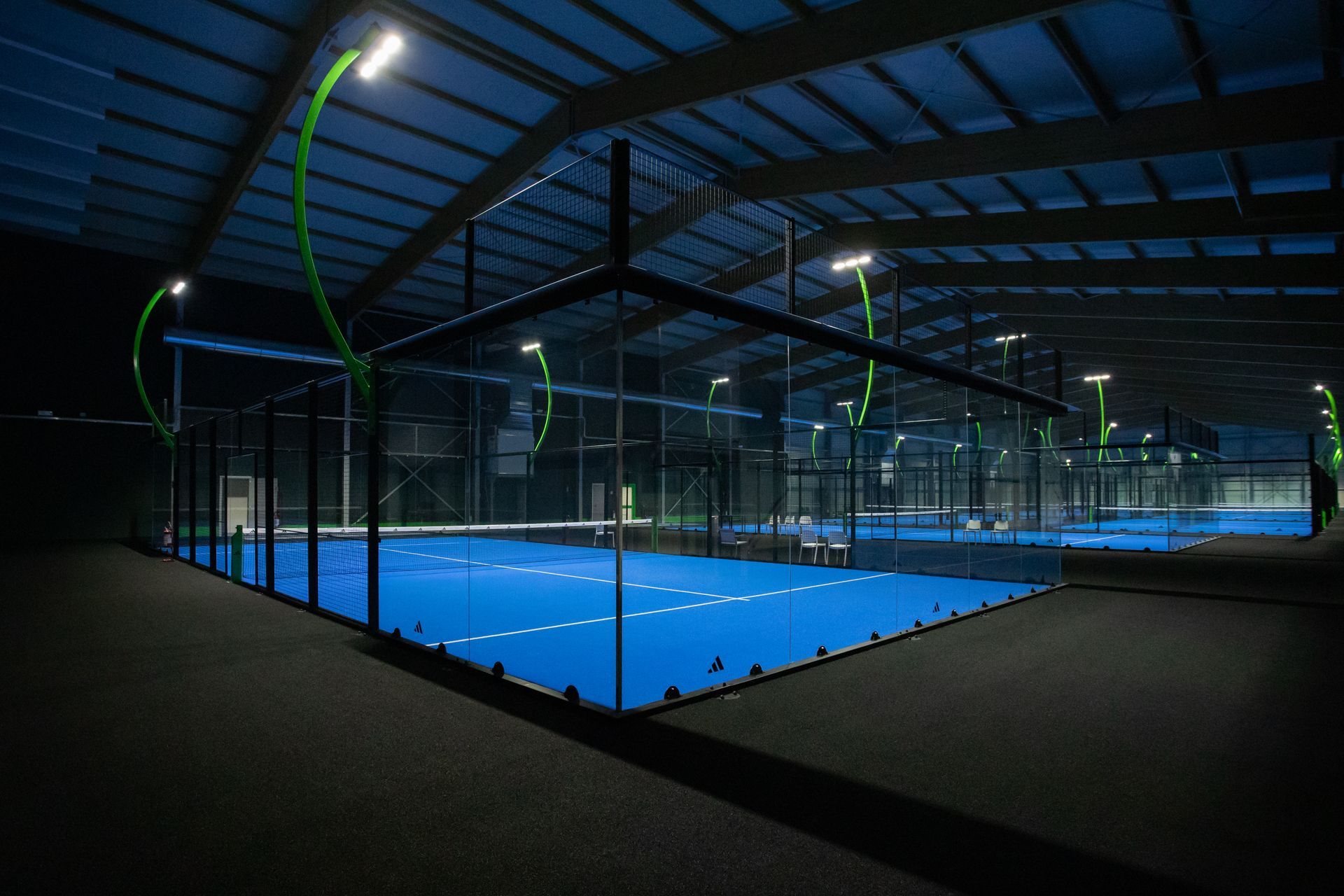 Campo da padel indoor, pavimento blu, rete nera, illuminazione intensa dall'alto.