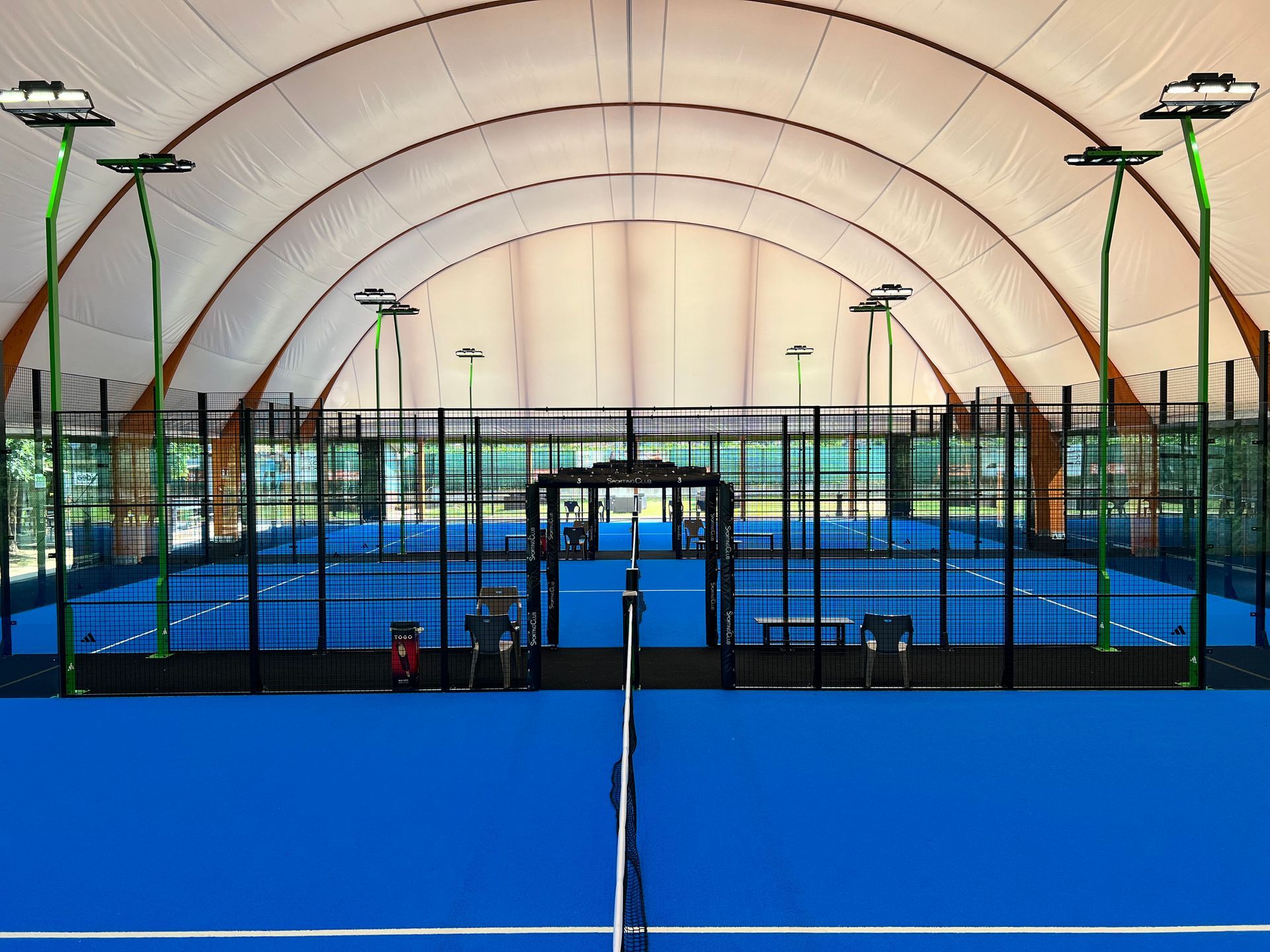 Campi da padel blu all'interno di una struttura bianca a forma di cupola, recinzione nera, illuminazione dall'alto.
