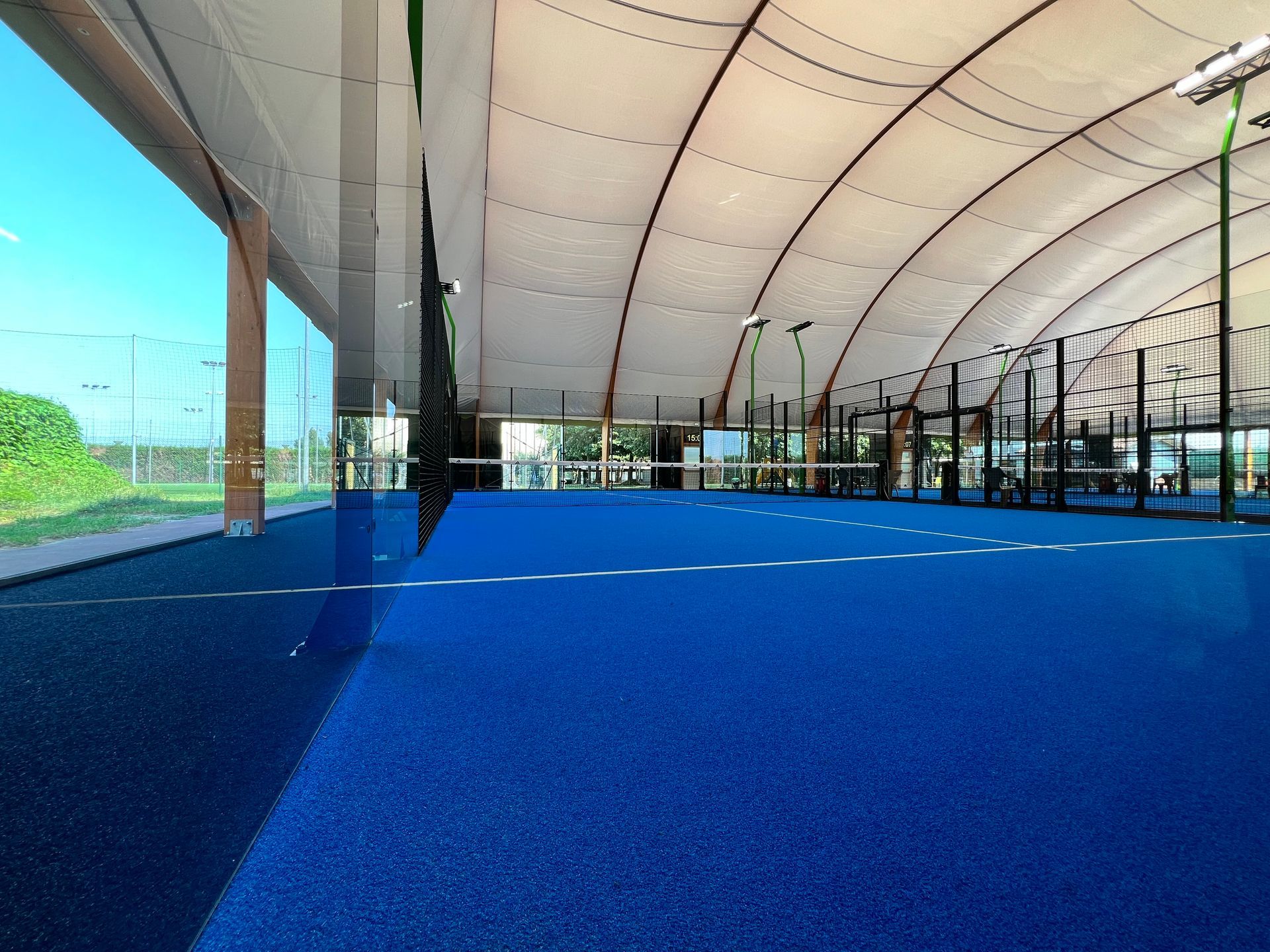 Campo da padel blu all'interno di una grande struttura a baldacchino bianco, con un'area verde 