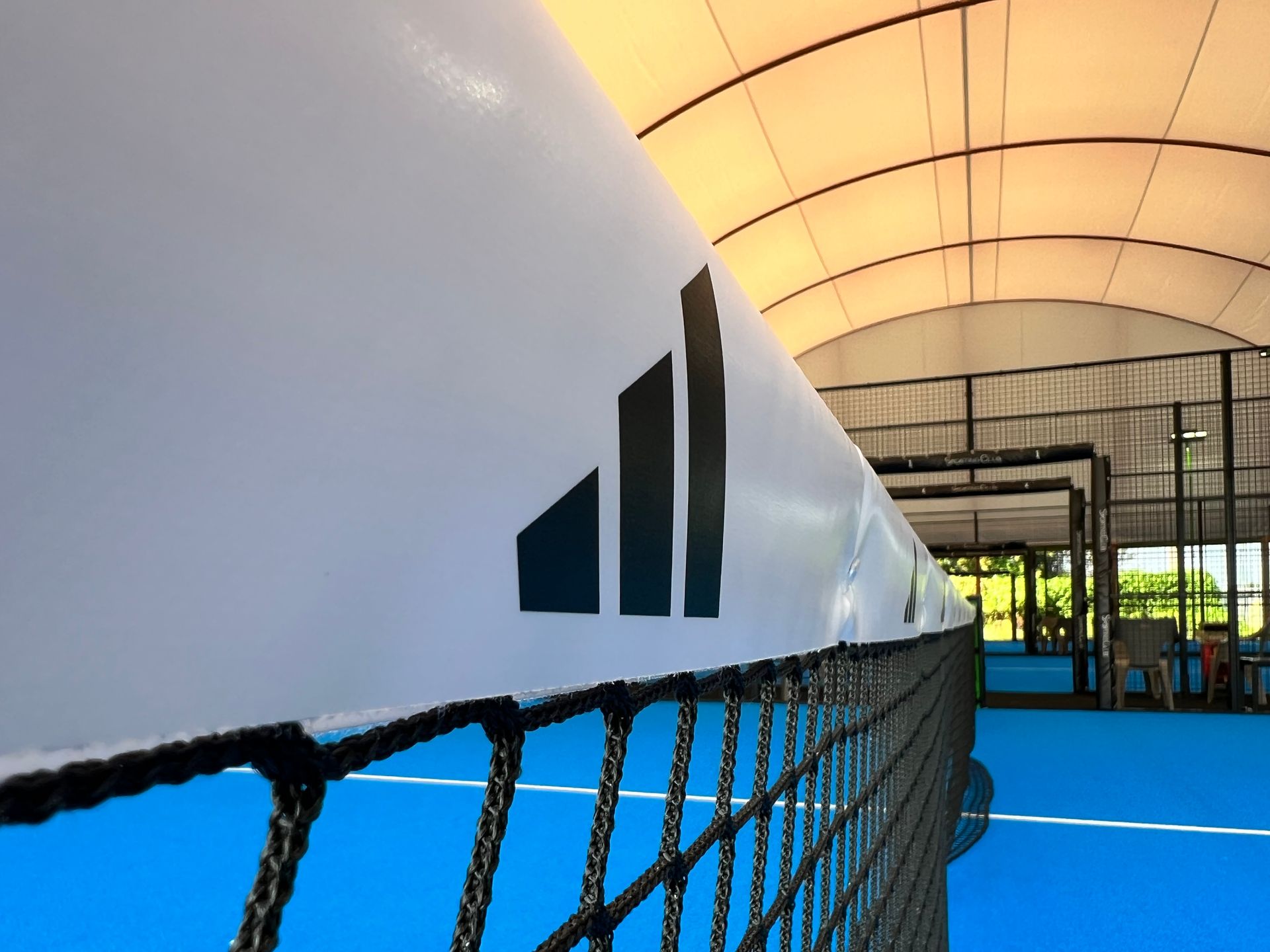 Primo piano di una rete da tennis con il logo Adidas. Campo blu, striscione bianco e ambientazione interna.