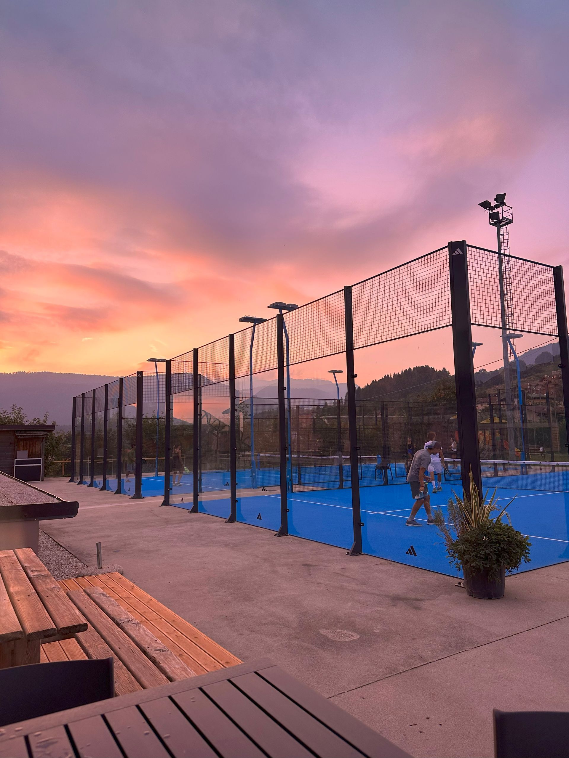Campo da padel al tramonto con giocatori sul campo blu, montagne e cielo colorato.