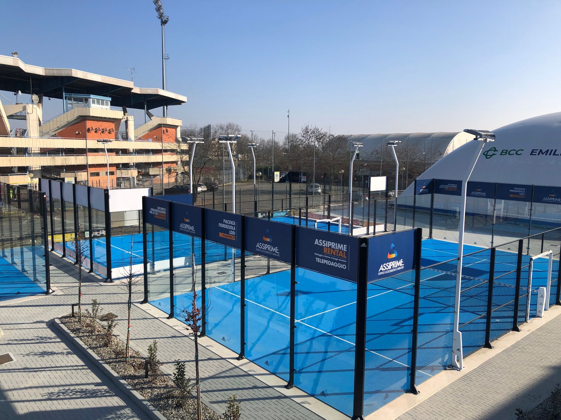 Diversi campi da padel blu all'aperto con uno stadio sullo sfondo in una giornata di sole.