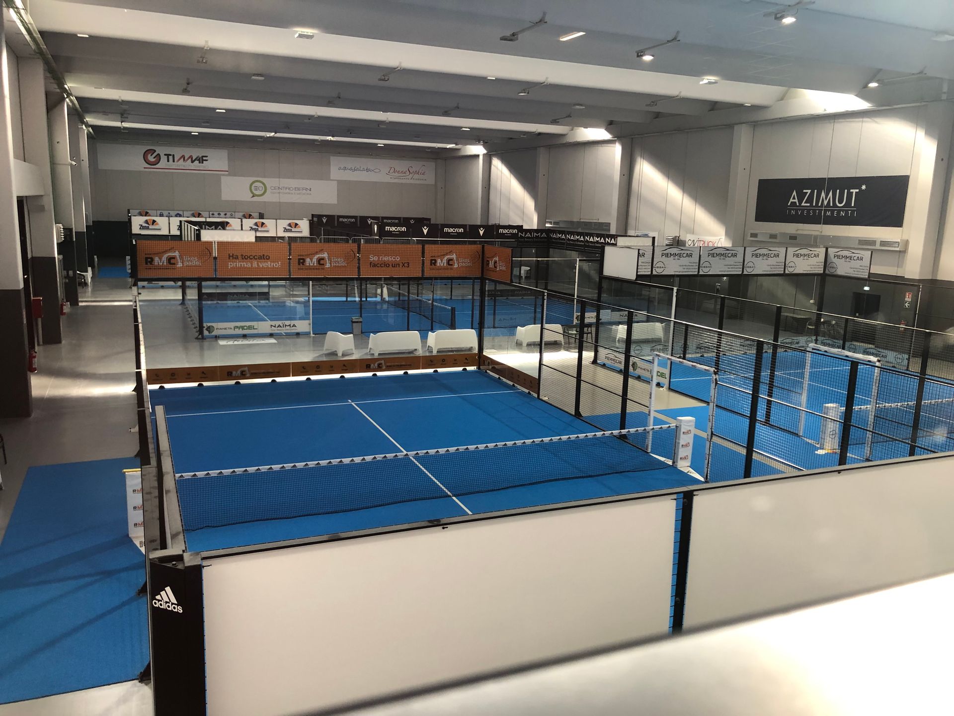 Campo da padel indoor con campi blu, circondato da vetrate e pareti bianche.