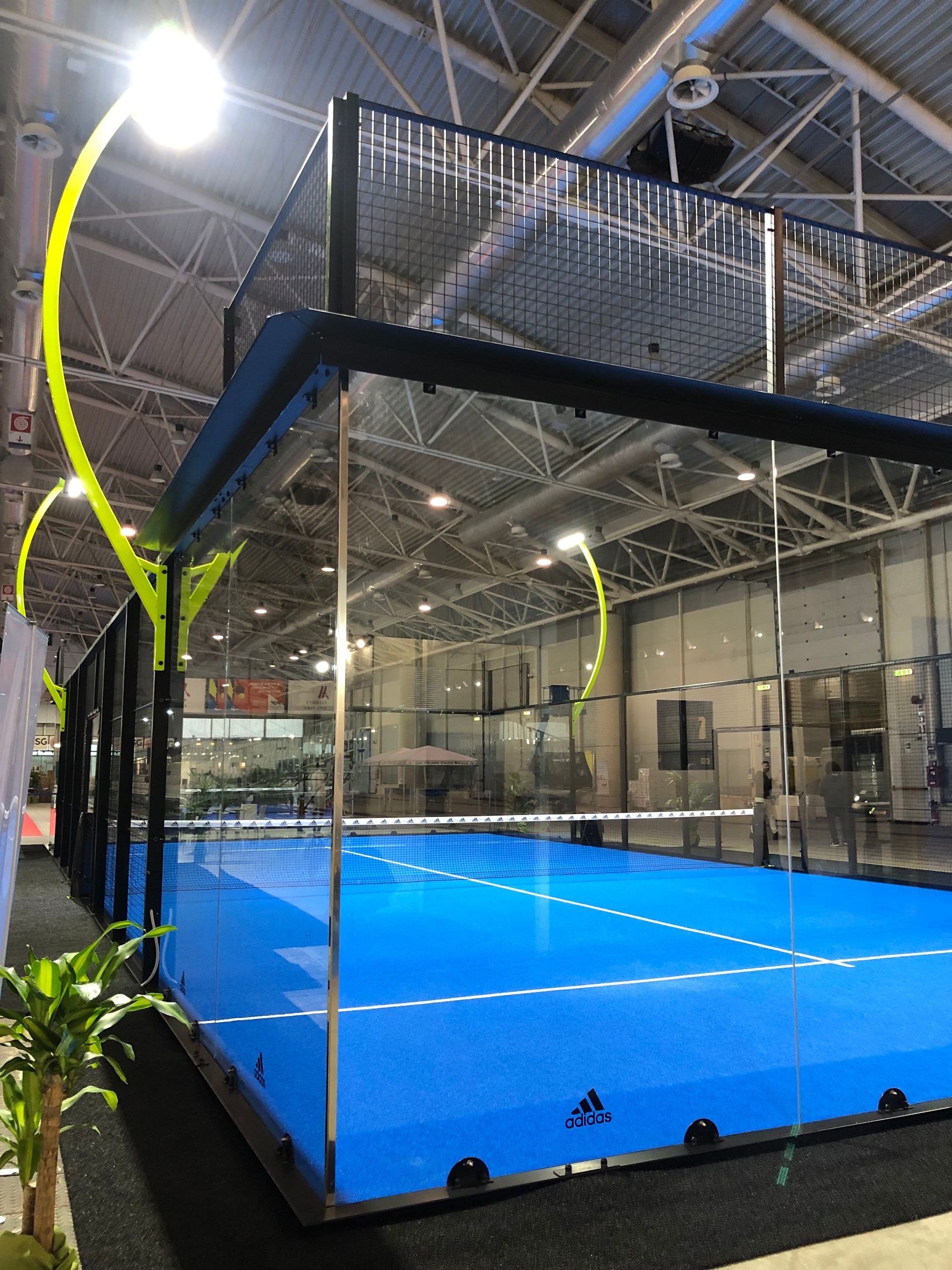 Campo da padel blu al coperto con pareti in vetro, rete nera e illuminazione dall'alto.
