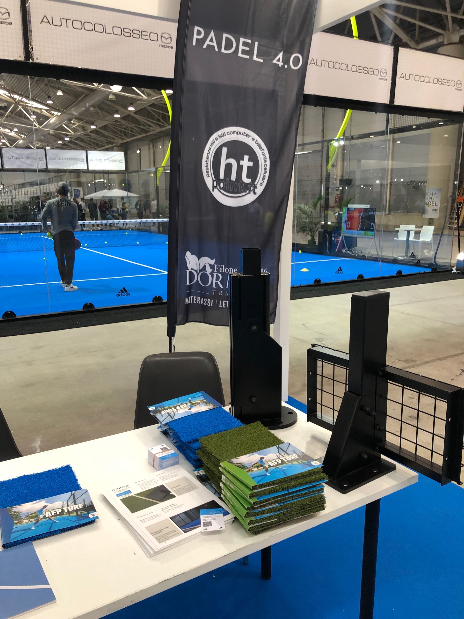 Tavolo con brochure e materiale promozionale per Padel 4.0. Sullo sfondo si vede un campo da tennis.