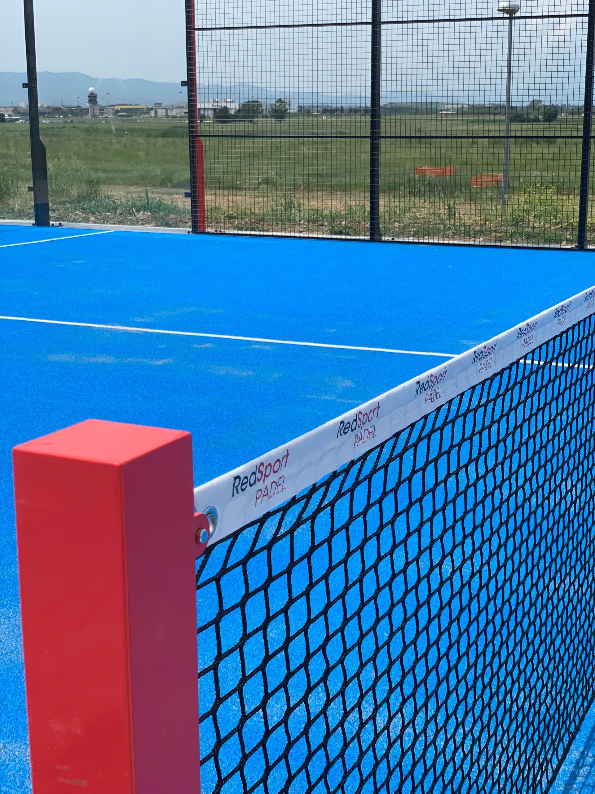Campo da padel blu con pali e rete rossi; ambiente esterno.