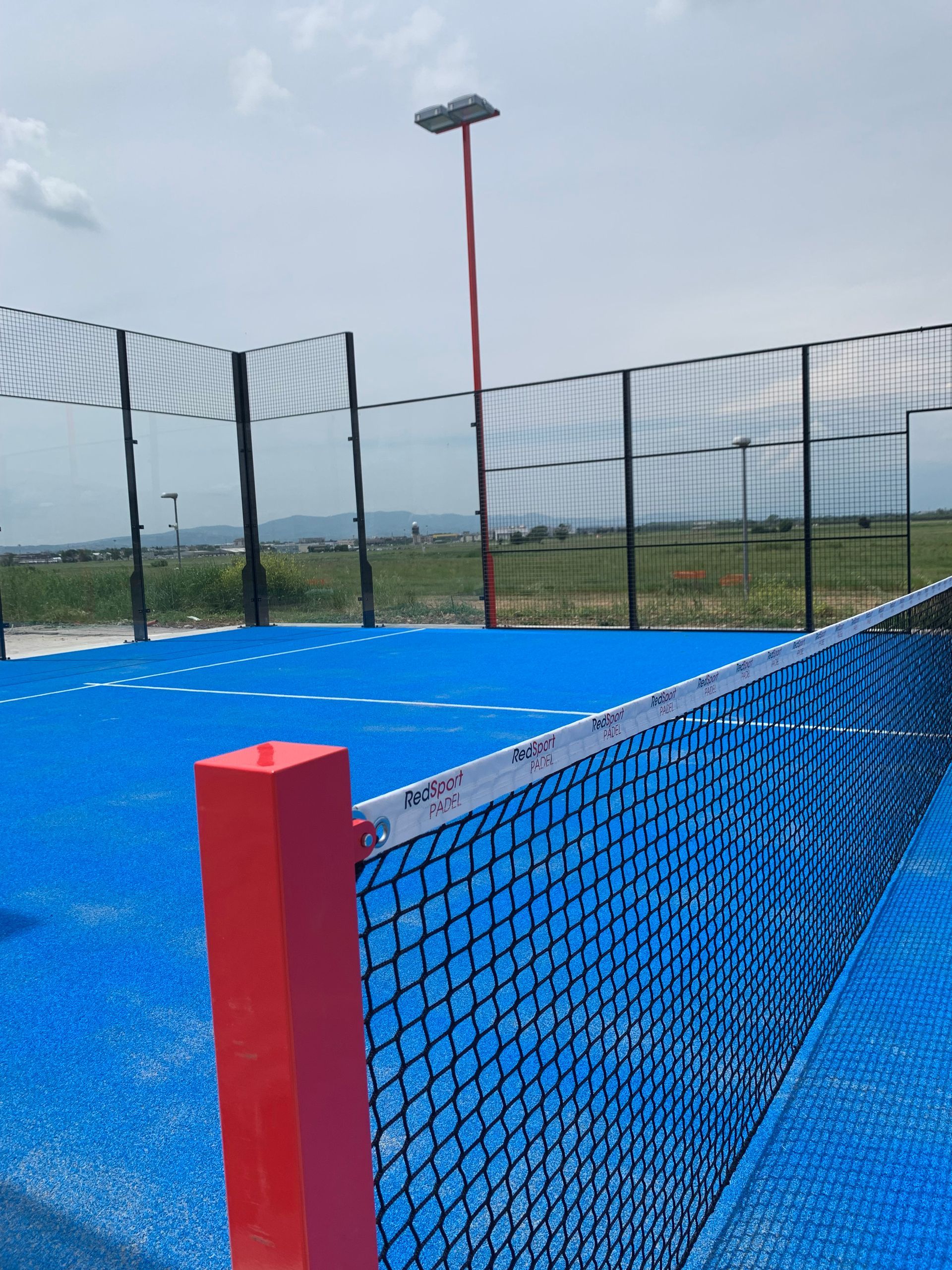 Campo da paddle tennis blu con pali della rete rossi, rete, recinzione e un alto palo della luce rosso.