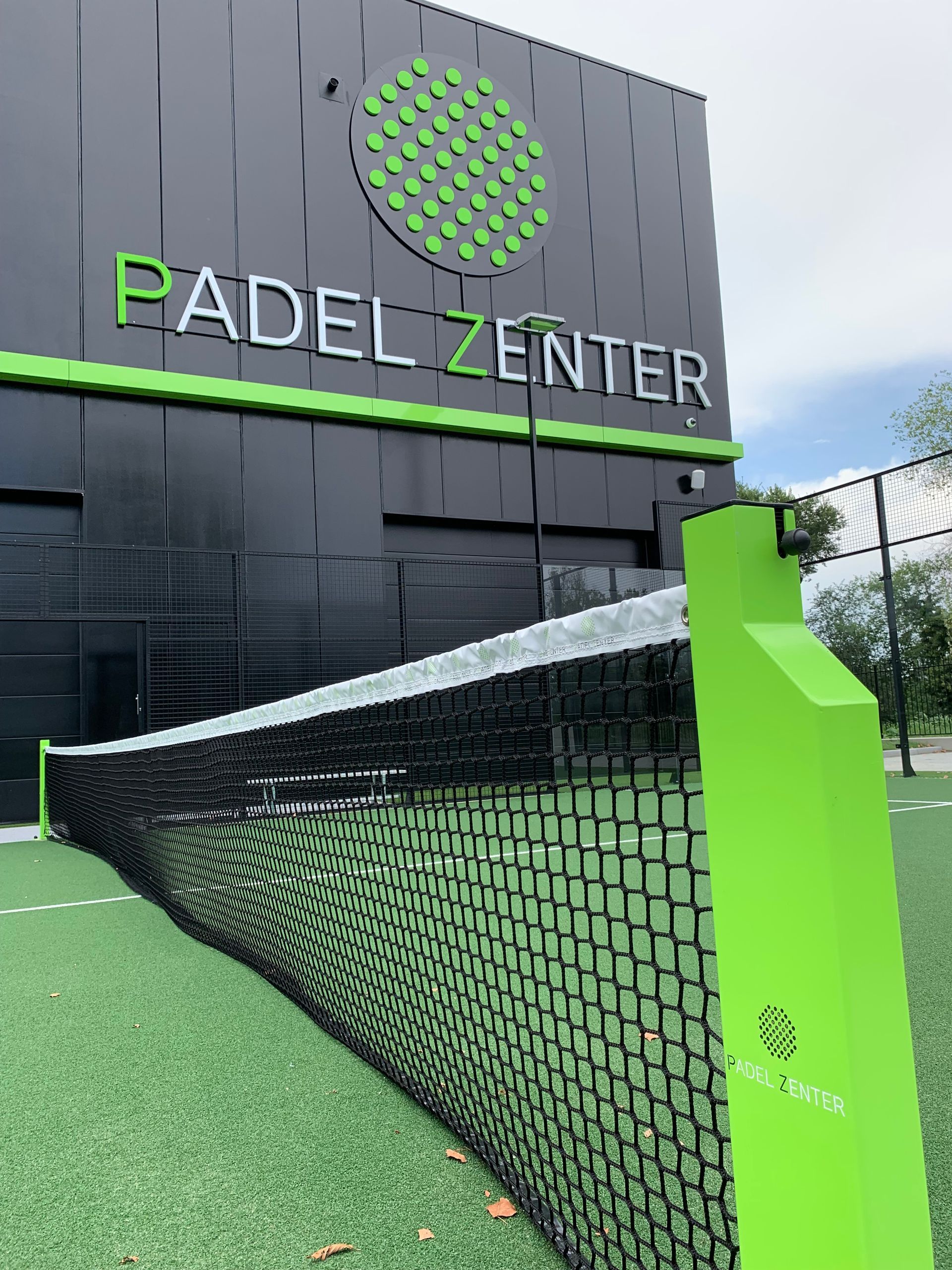 Insegna Padel Zenter su un edificio nero con dettagli verdi. In primo piano una rete da tennis.