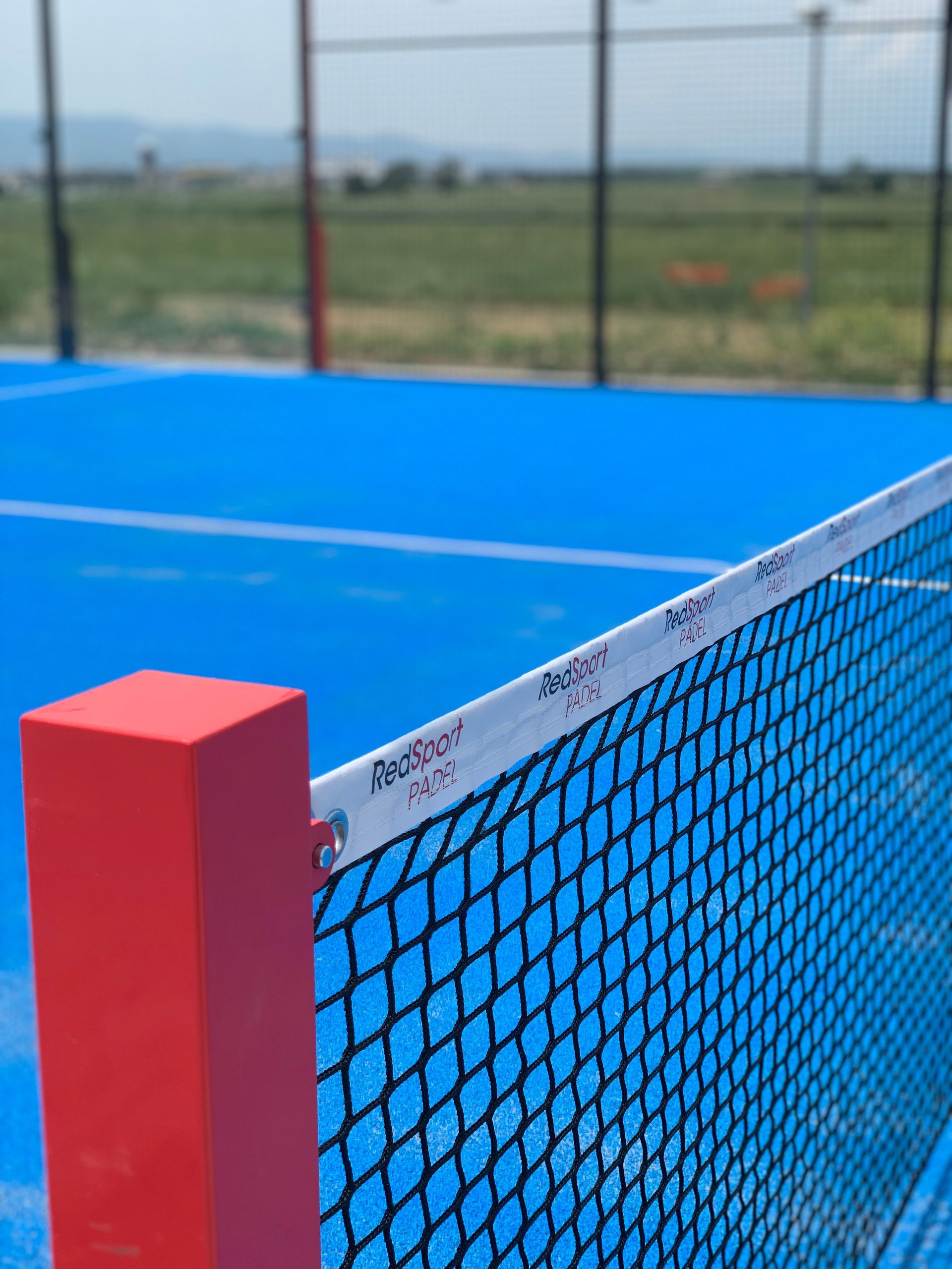 Campo da pickleball blu con rete e palo rossi; ambiente esterno soleggiato.