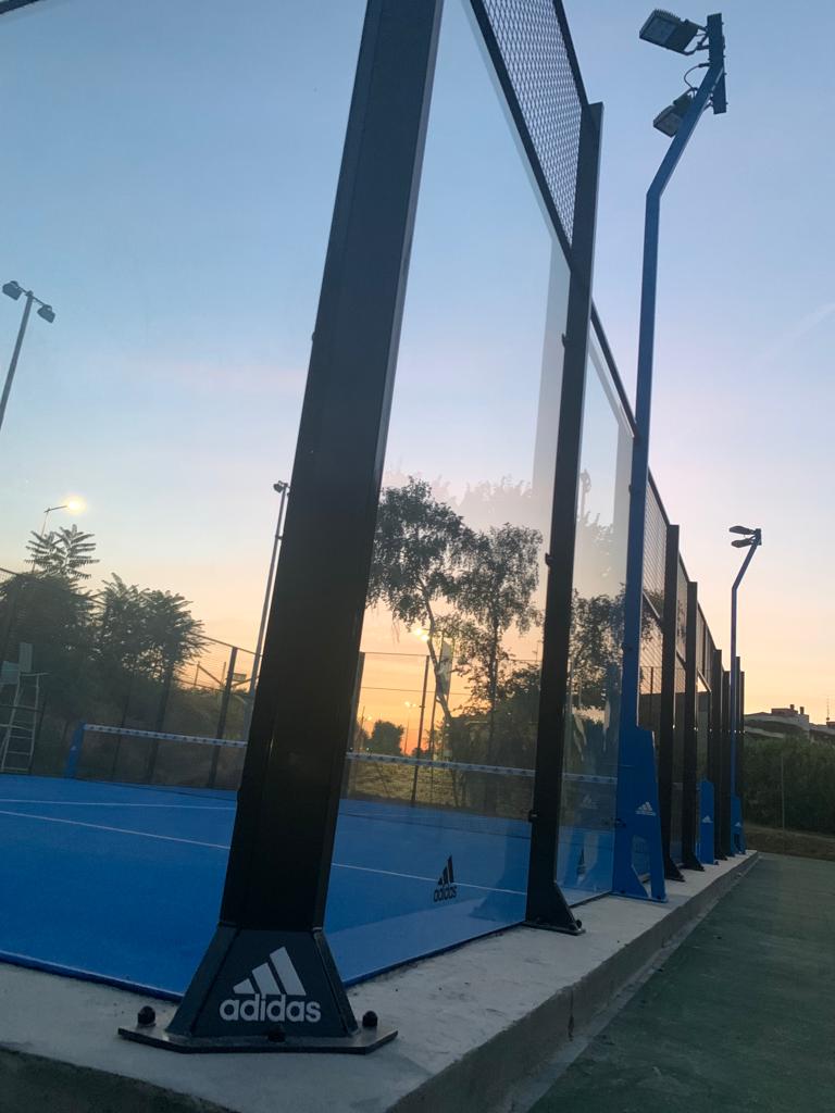 Campo da padel blu con pareti in vetro, supporti neri e logo Adidas, sotto un cielo serale.