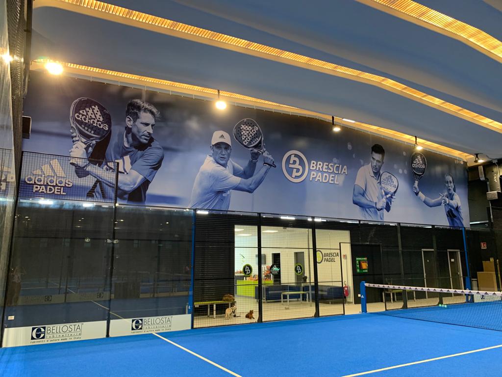 Campo da padel blu al coperto con uno striscione raffigurante giocatori; persone sullo sfondo.