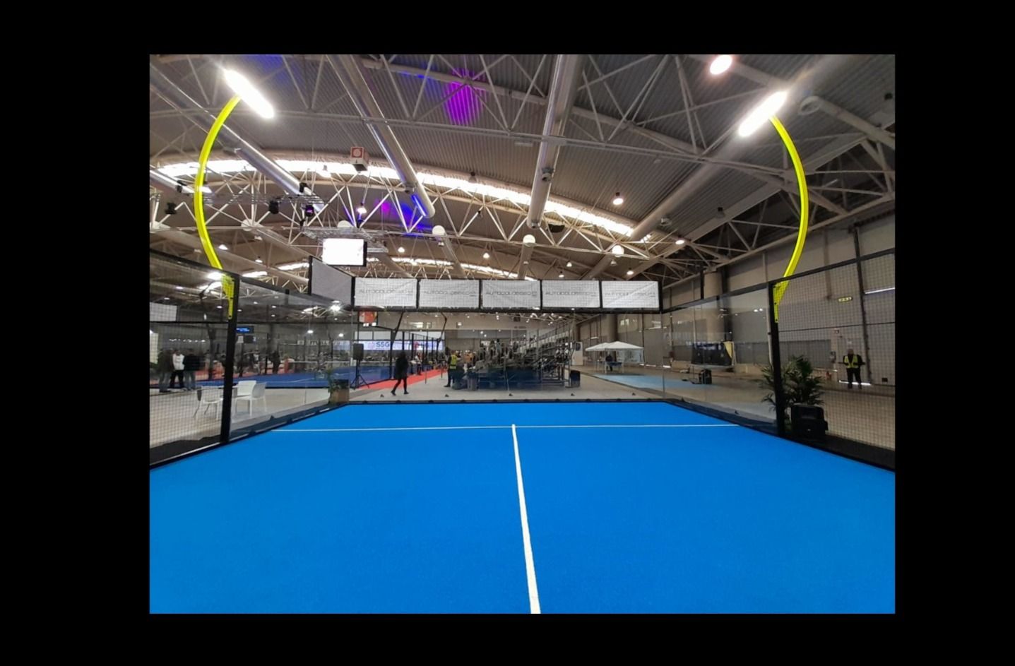 Campo da padel coperto, superficie blu, illuminato da lampade sospese. Giocatori visibili.