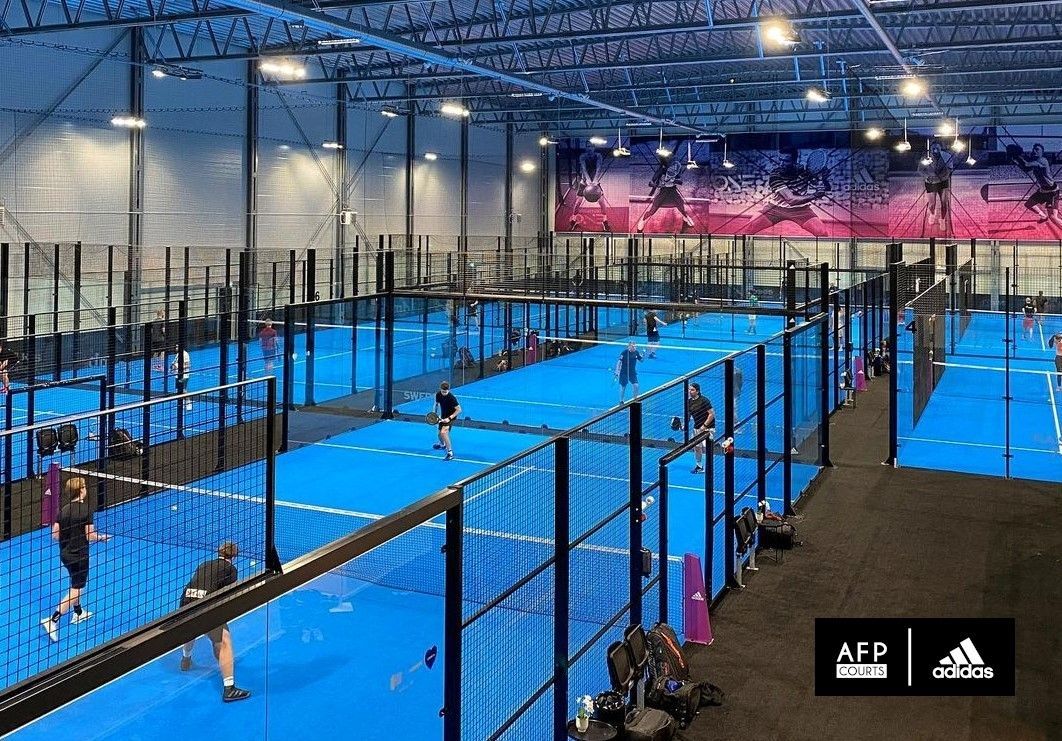 Campi da padel indoor con giocatori. Campi blu, recinzione nera, illuminazione dall'alto.