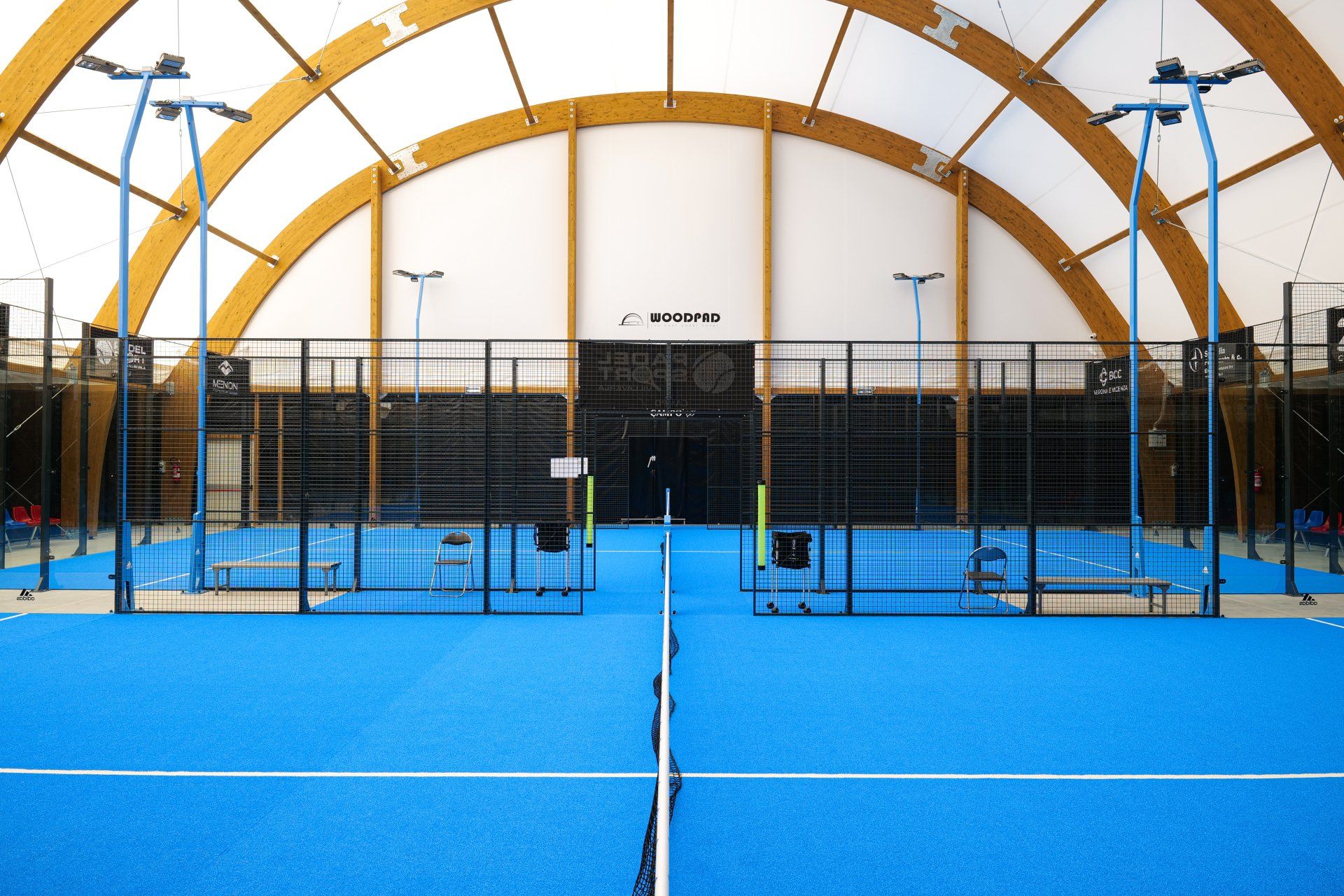 Campo da padel al coperto, superficie blu, recinzione nera, soffitto in legno ad arco, illuminazione dall'alto.