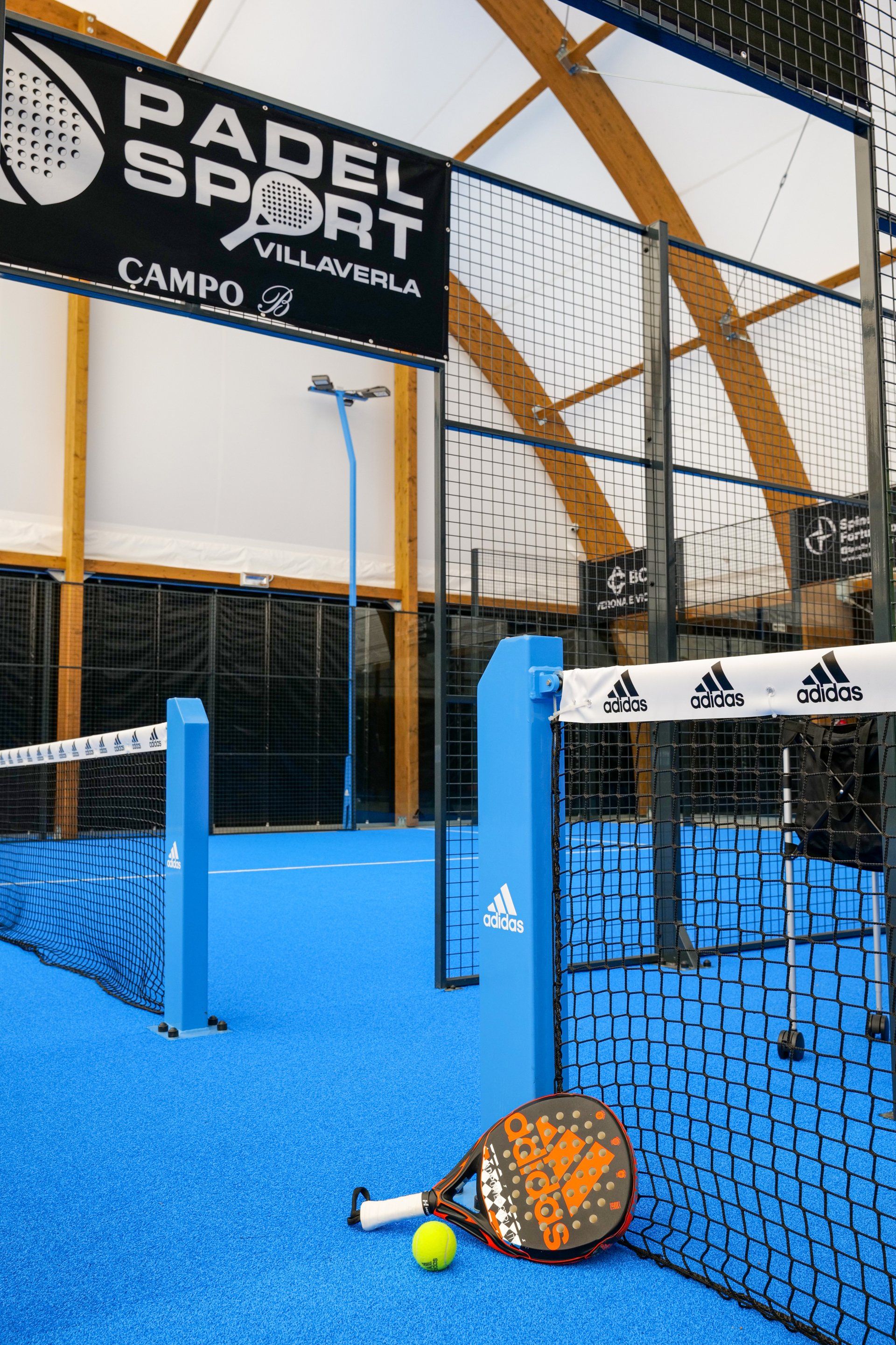 Campo da padel indoor, superficie blu, rete nera, logo Adidas, racchetta e pallina.