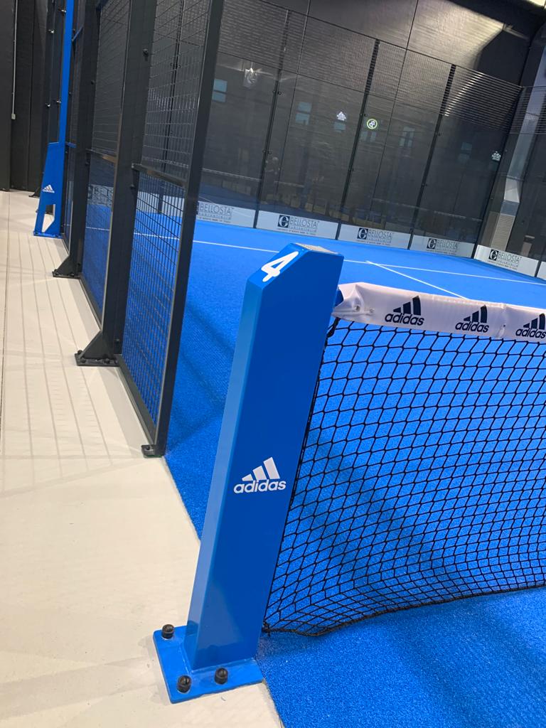 Campo da padel Adidas blu e nero con rete, logo sul palo, ambiente indoor.