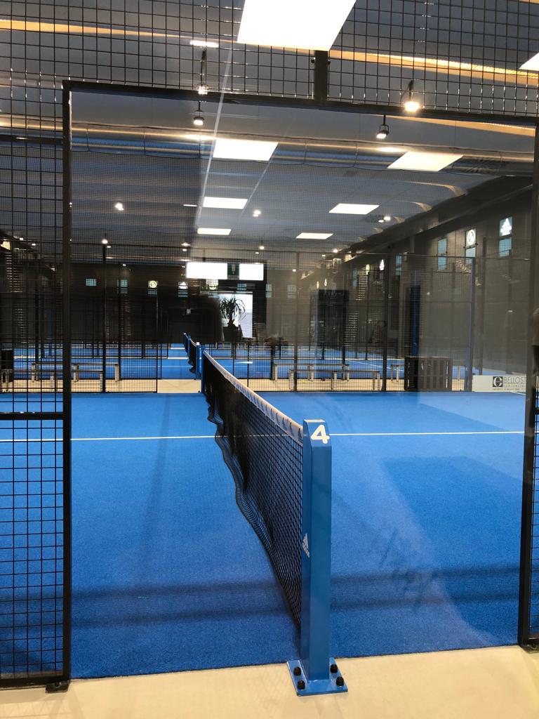 Campo da padel blu con rete nera, ambientazione interna, illuminazione dall'alto.