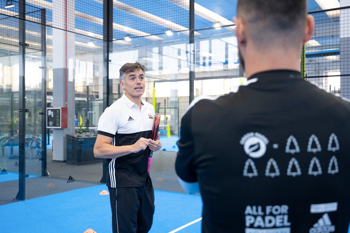 Un uomo con una camicia bianca istruisce una persona con una camicia nera su un campo da padel blu.